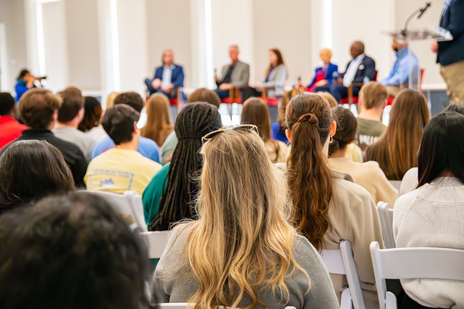 Audience members seated in white chairs attend a panel discussion with speakers at the front of a bright conference room.