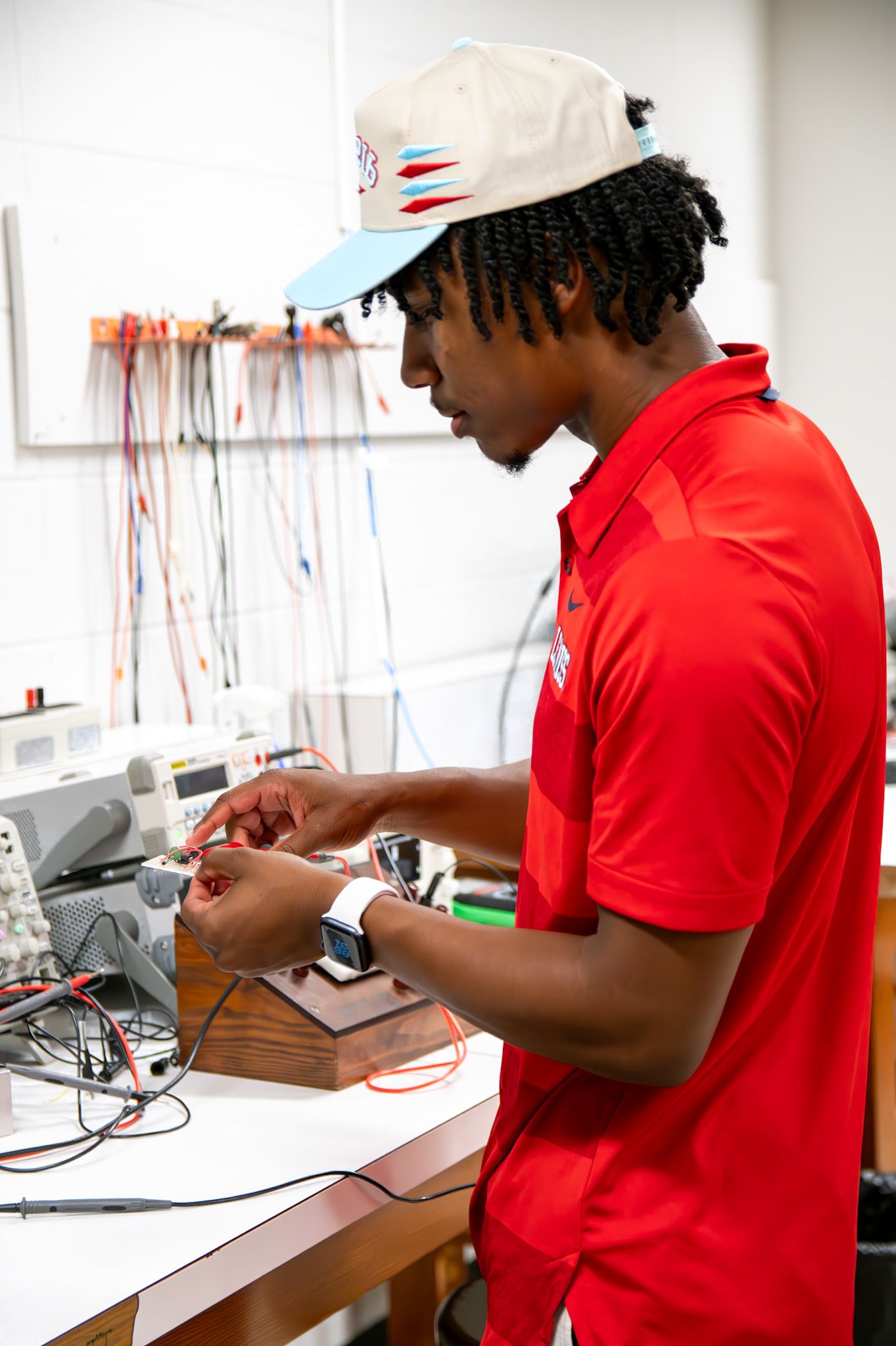 Young man in red shirt and cap works with electronic components at a lab workbench with wires and equipment.