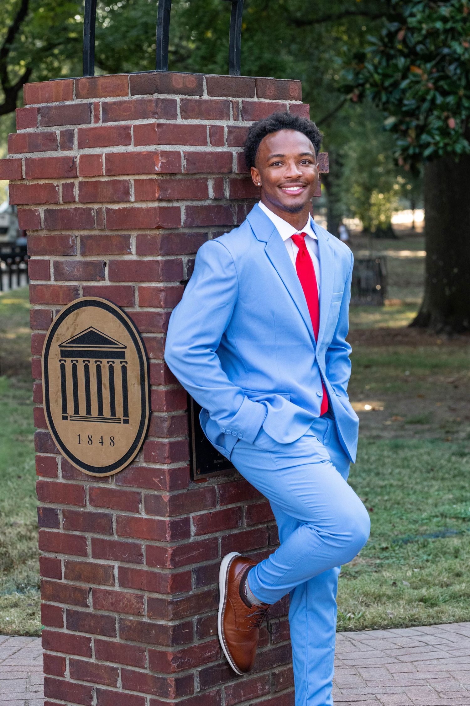 Young man in light blue suit and red tie posing confidently by a brick pillar with 1848 university seal.