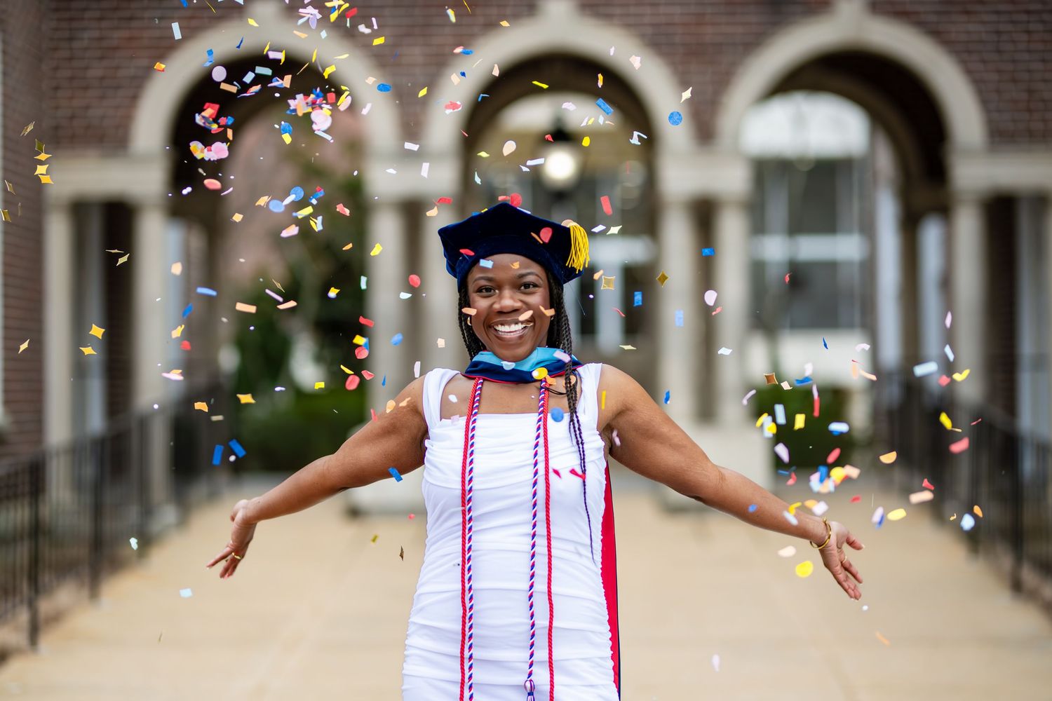 Graduate in cap and multicolored stole celebrates with arms outstretched as confetti falls in front of arched building.