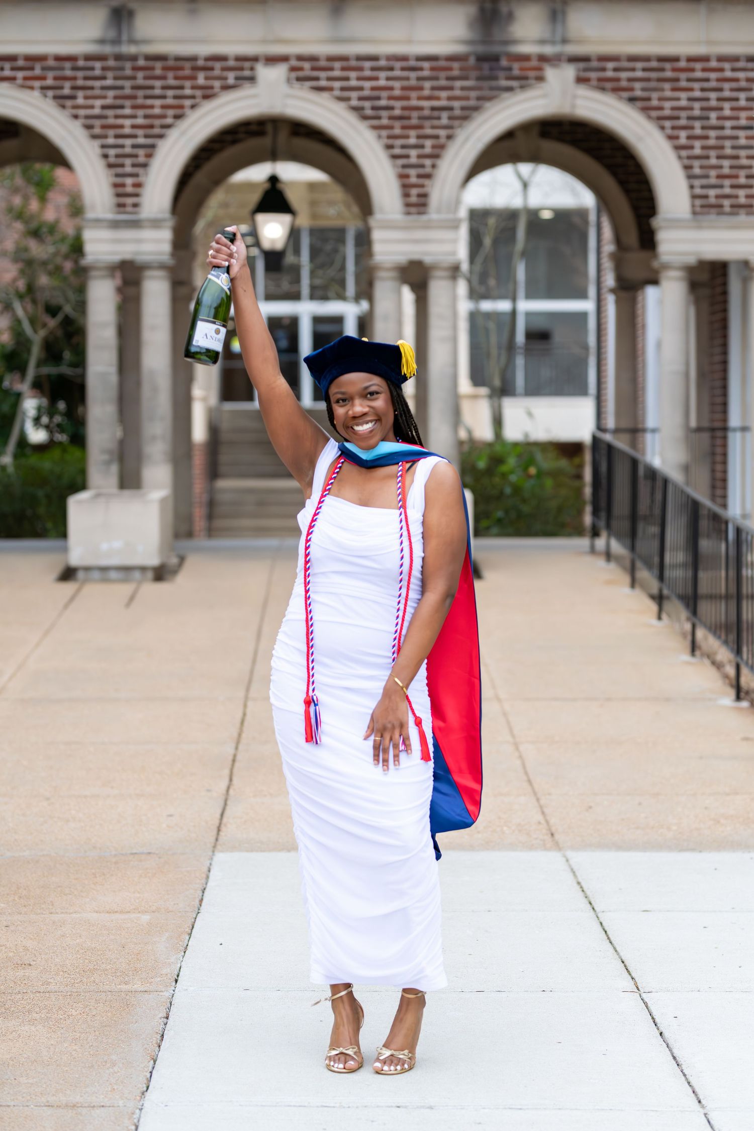 Graduate stands confidently on a brick building walkway wearing a white dress and graduation cap for commencement photos.