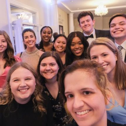 A large group of smiling young adults dressed in formal attire pose together for a selfie at an elegant event.