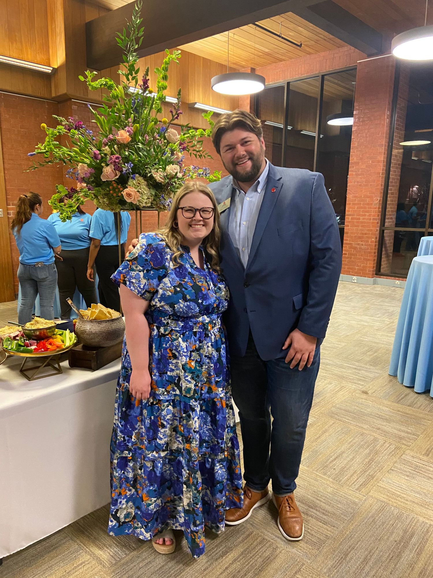 A smiling couple poses together at an indoor event, woman in floral blue dress and man in blue blazer.