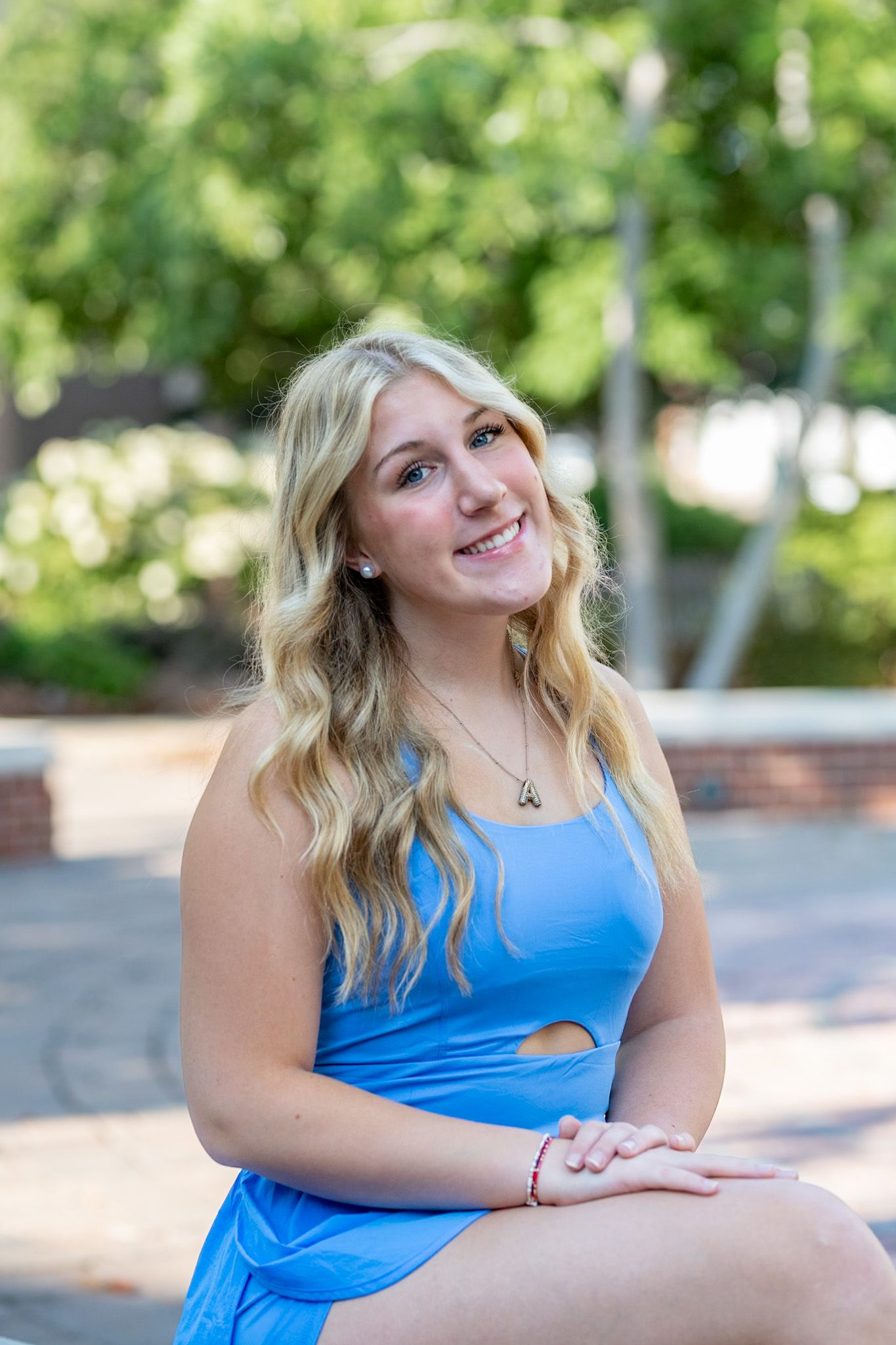 Young blonde woman in a blue dress sitting outdoors, smiling cheerfully in a lush green garden setting.