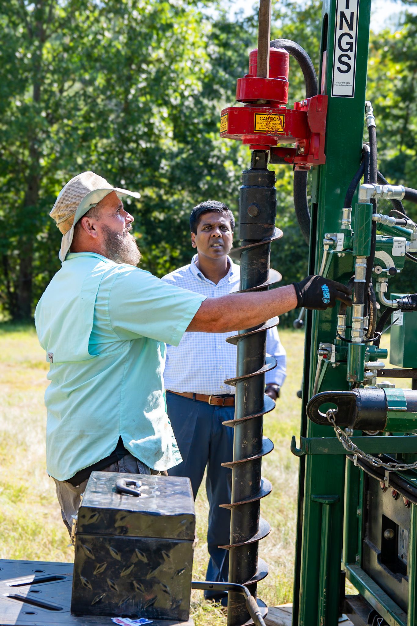 Two men operating a green drilling rig outdoors, one in a cap and gloves, another in a dress shirt, surrounded by trees.