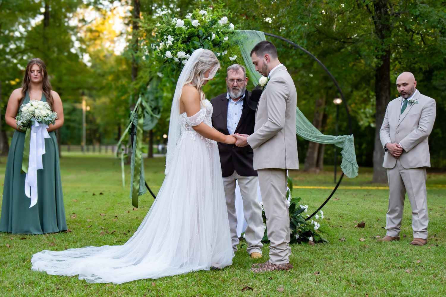 A wedding ceremony takes place outdoors on a lush green lawn with an arch and attendants in soft blue and grey attire.