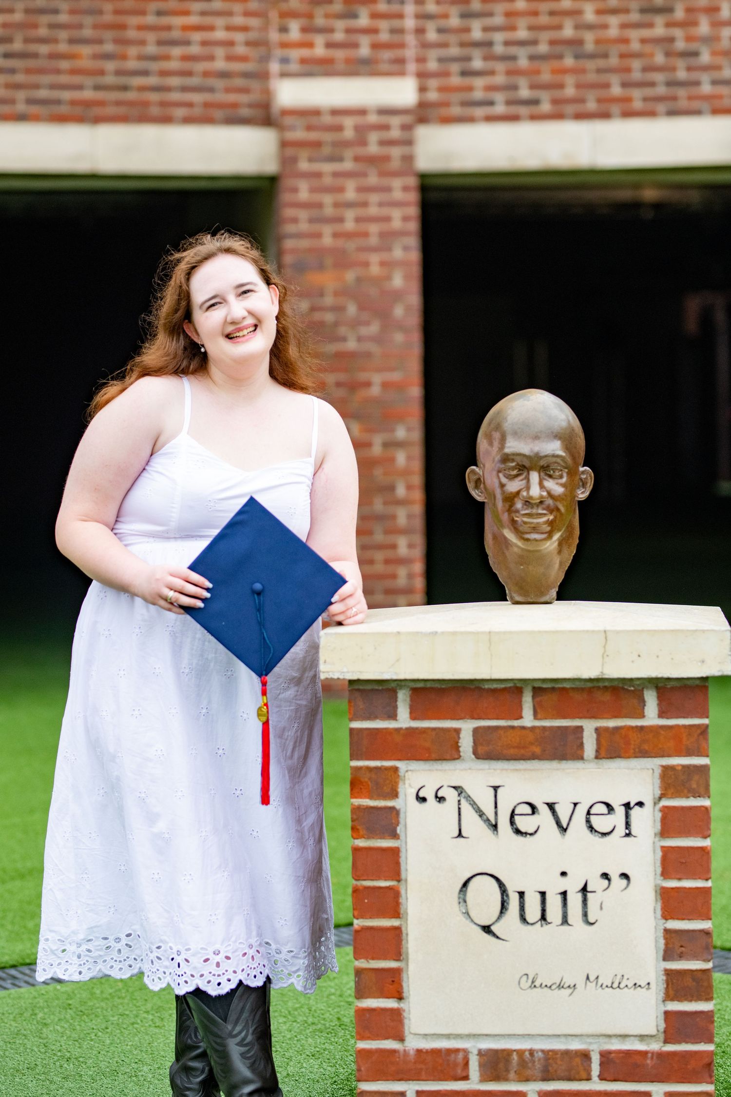 Graduate in white dress poses with diploma next to bronze sculpture and 'Never Quit' inspirational brick wall.