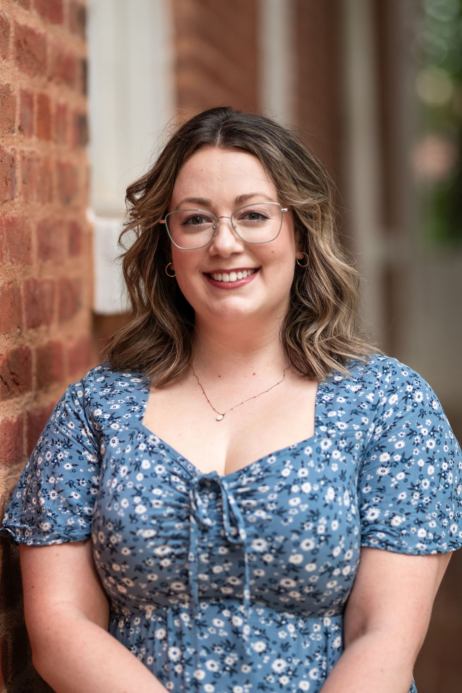 Portrait photos showing person in blue floral dress with shoulder-length hair and glasses against brick backdrop.
