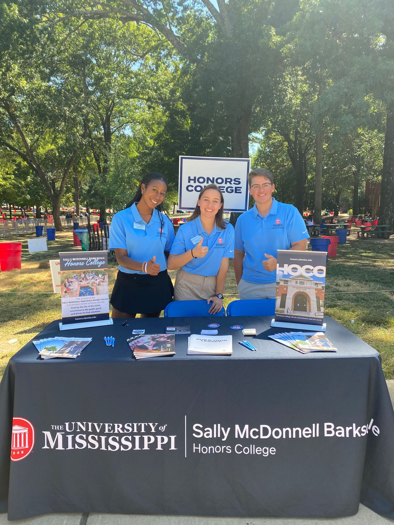 Three students in blue shirts smile at the University of Mississippi Sally McDonnell Barksdale Honors College outdoor booth.