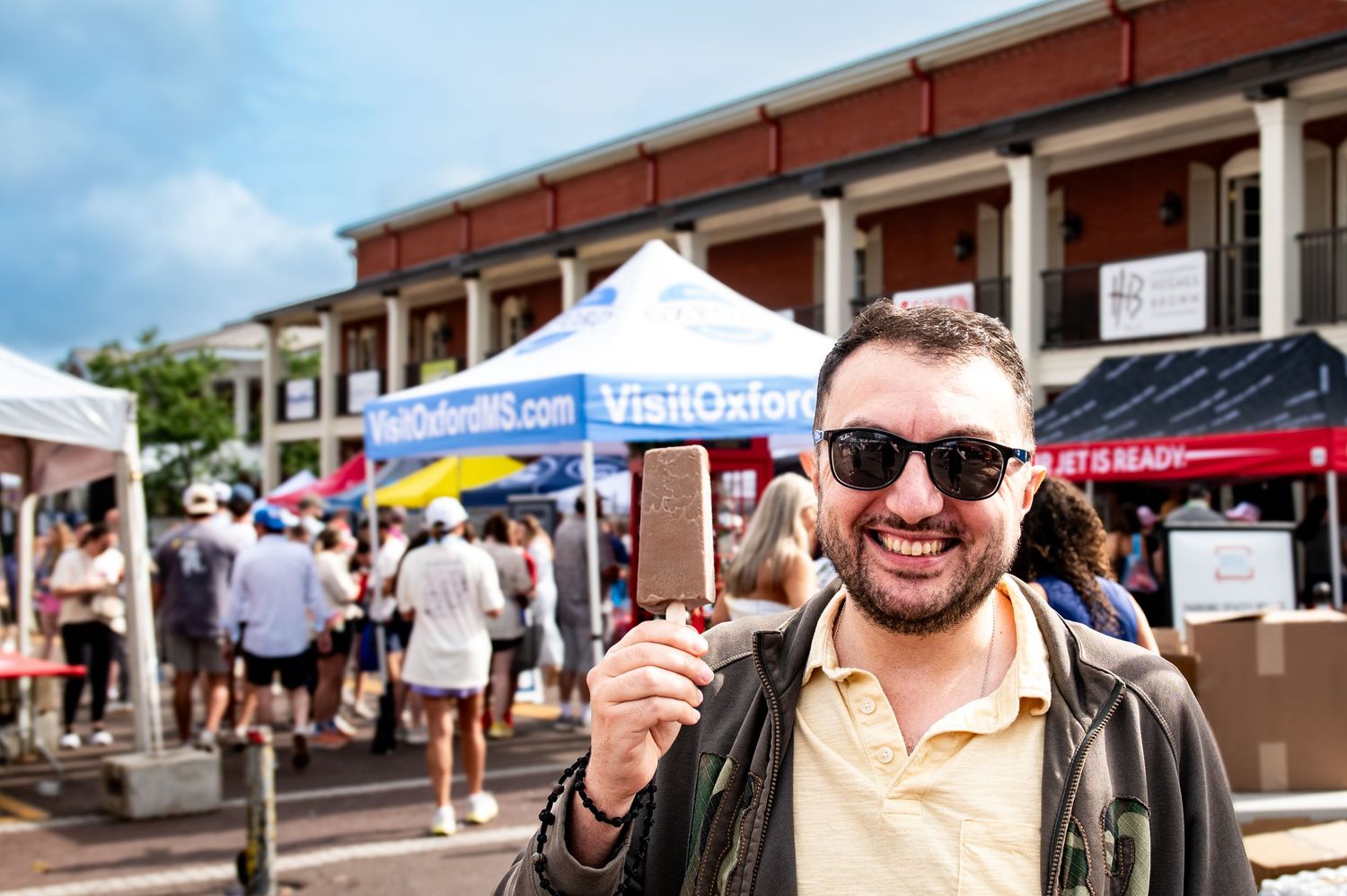 Man holding smartphone at Visit Oxford MS outdoor market event with colorful tents and crowd in background.