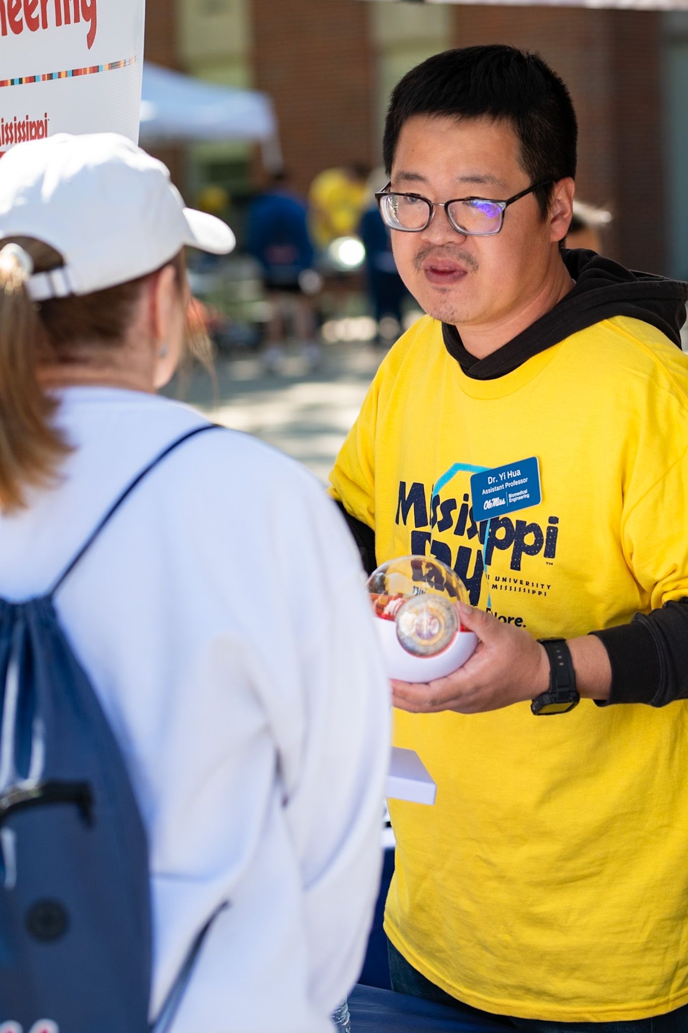 Mississippi State University staff member in yellow shirt engages with visitor at outdoor campus event.