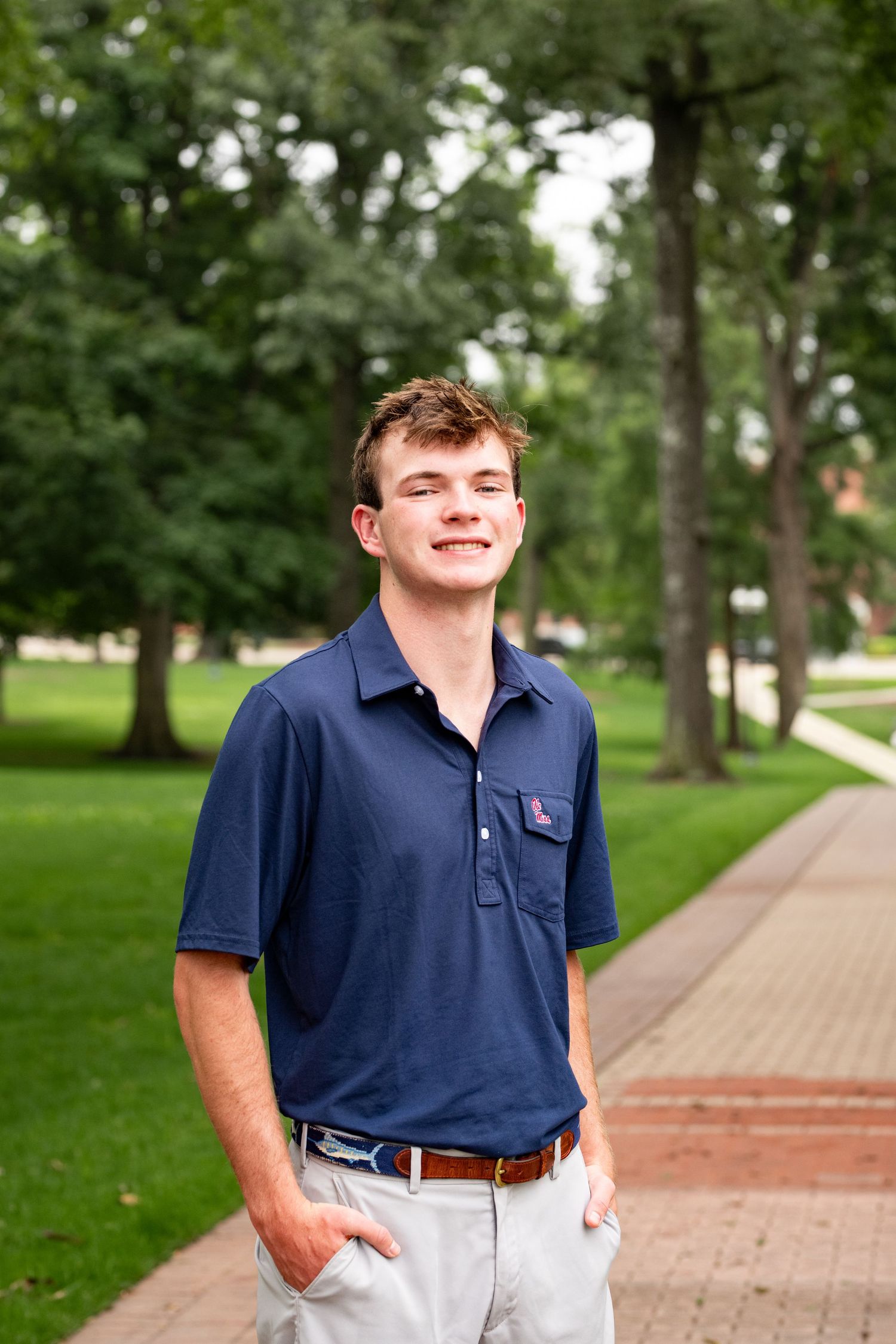 Smiling young man in navy polo shirt standing on a tree-lined campus walkway with hands in pockets.