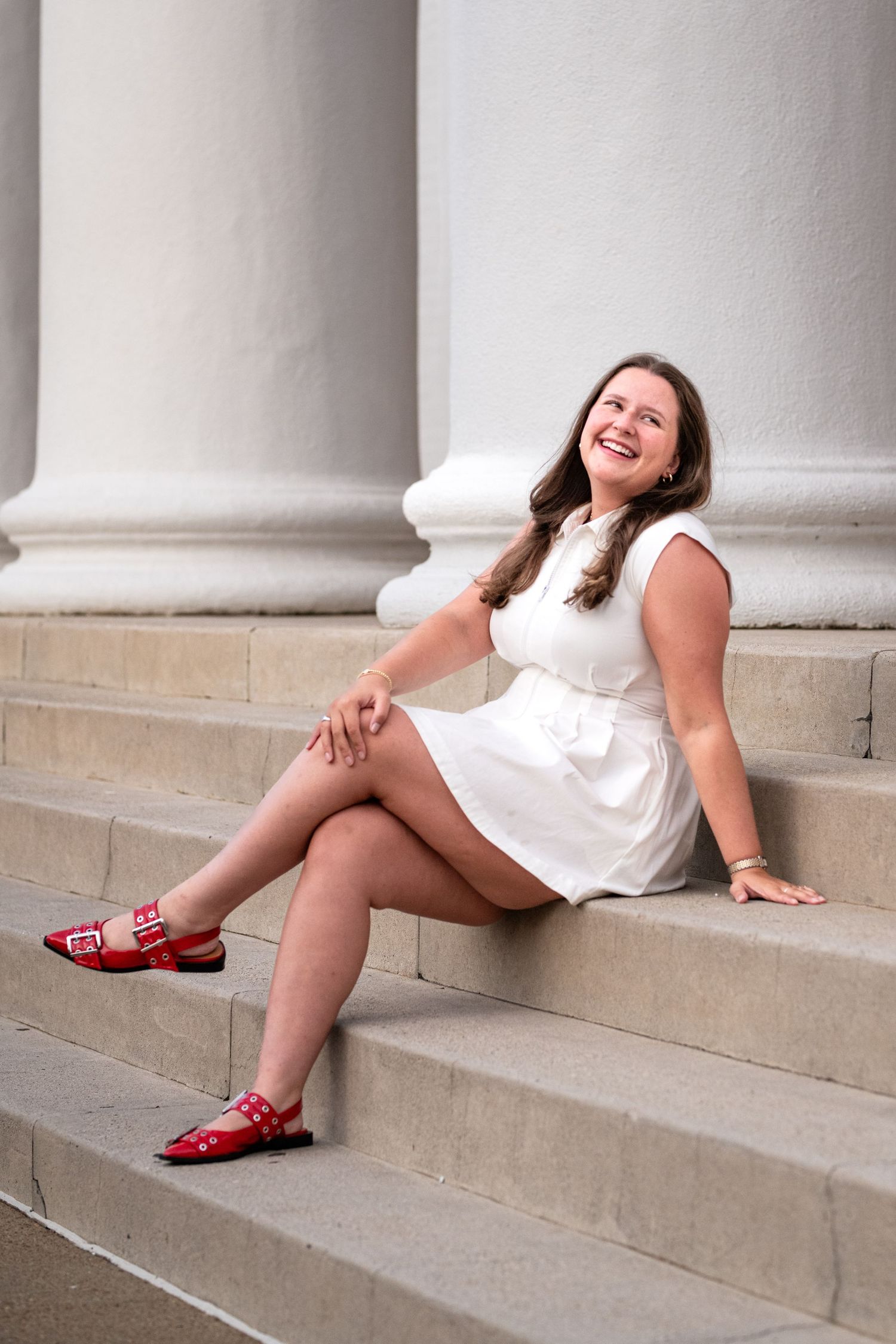 Young woman in white dress and red shoes sitting on stone steps near classical columns, smiling joyfully.