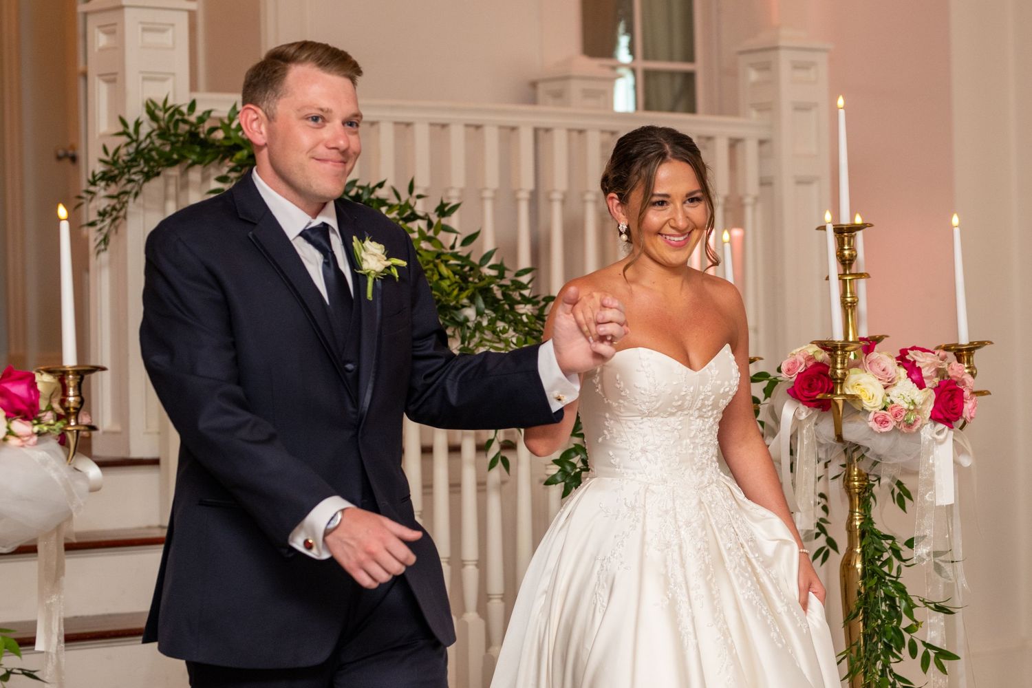 A bride and groom celebrate during their wedding ceremony in an elegant white interior with floral decorations and candles.