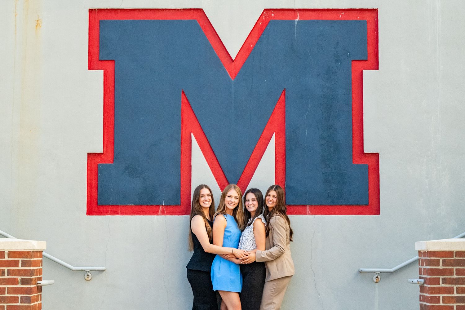 Four young women pose together in front of a large red and blue letter M on a gray wall.