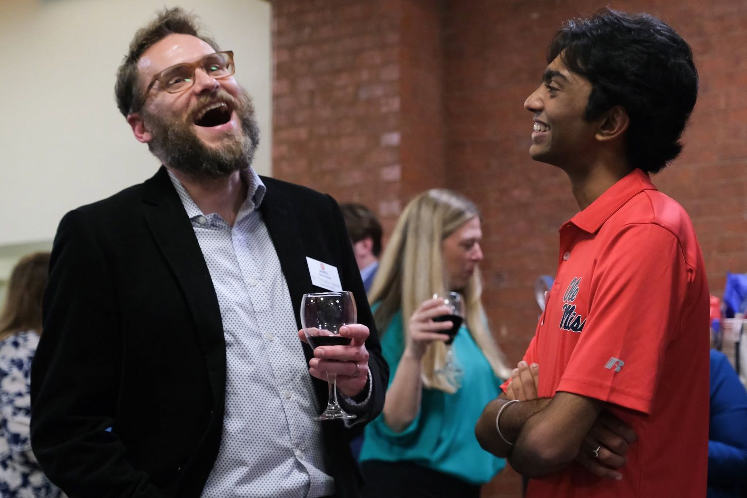 Two men laughing together at a networking event, one holding a glass of wine.