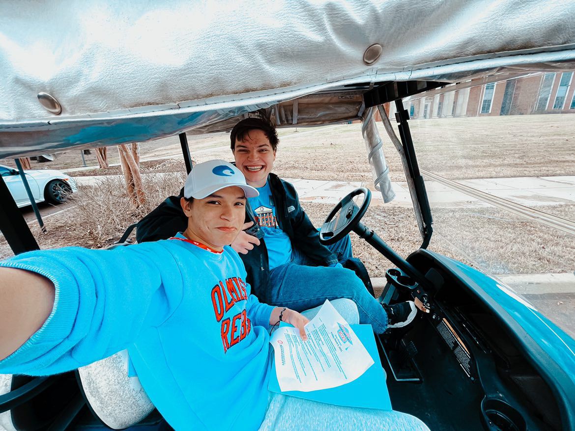 Two people in blue shirts smile for a selfie while riding in a golf cart outdoors.