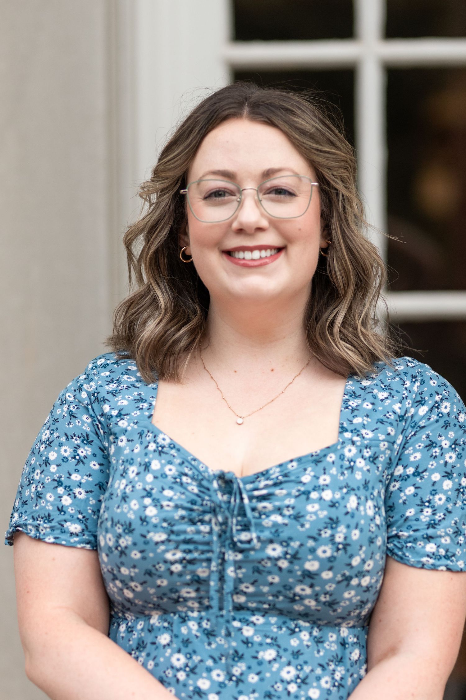 A woman in a blue floral dress and glasses smiles warmly against a white window backdrop.