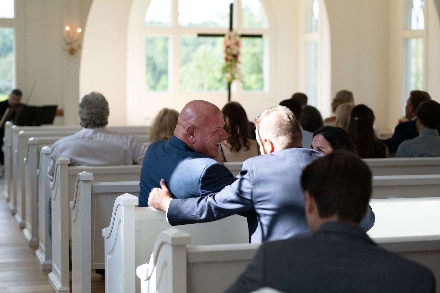 Two people embrace emotionally in a white church pew while other guests are seated during a ceremony.