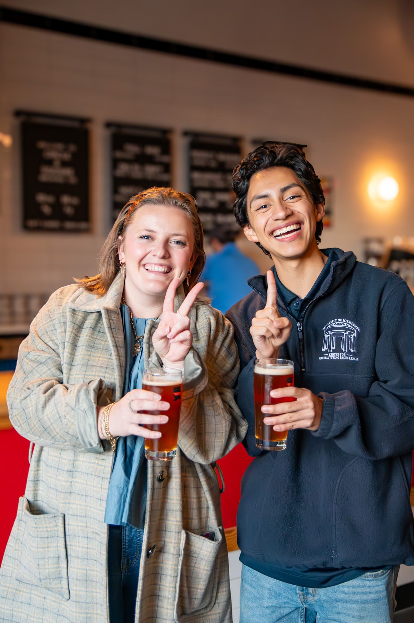Two smiling friends holding amber beers and making hand gestures at a brewery taproom.