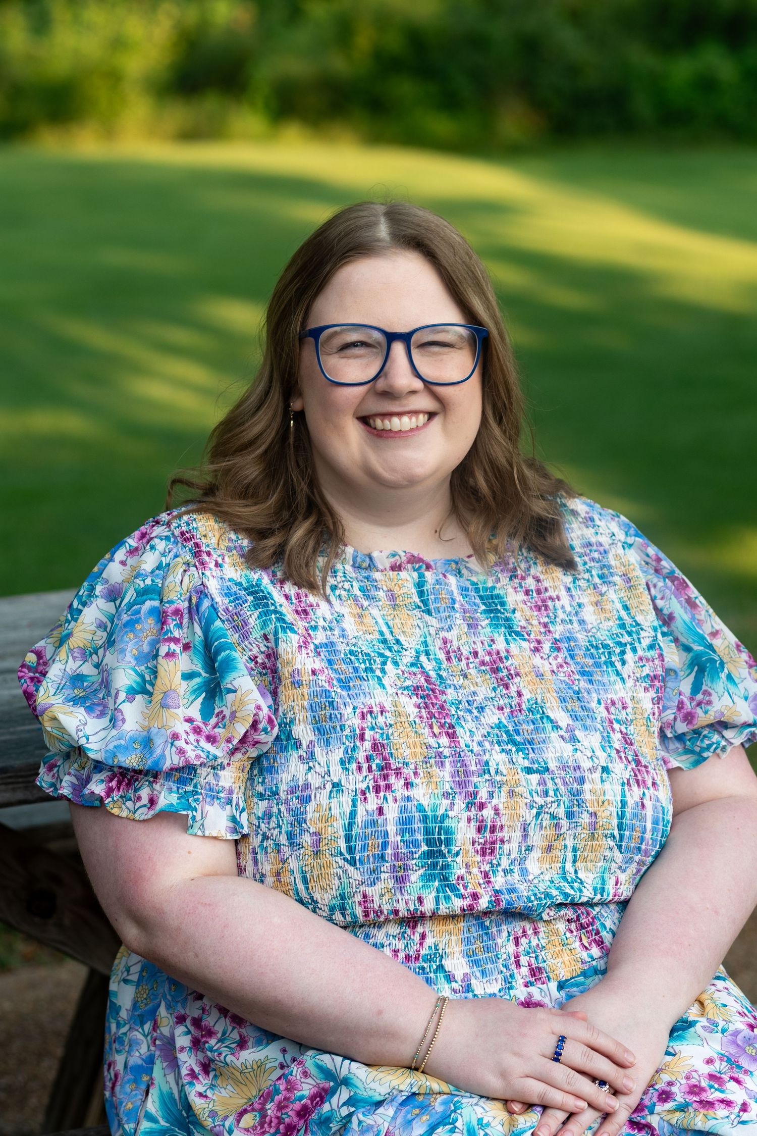 Smiling woman with blue glasses wearing a colorful floral dress, seated outdoors on a bench with green grass background.