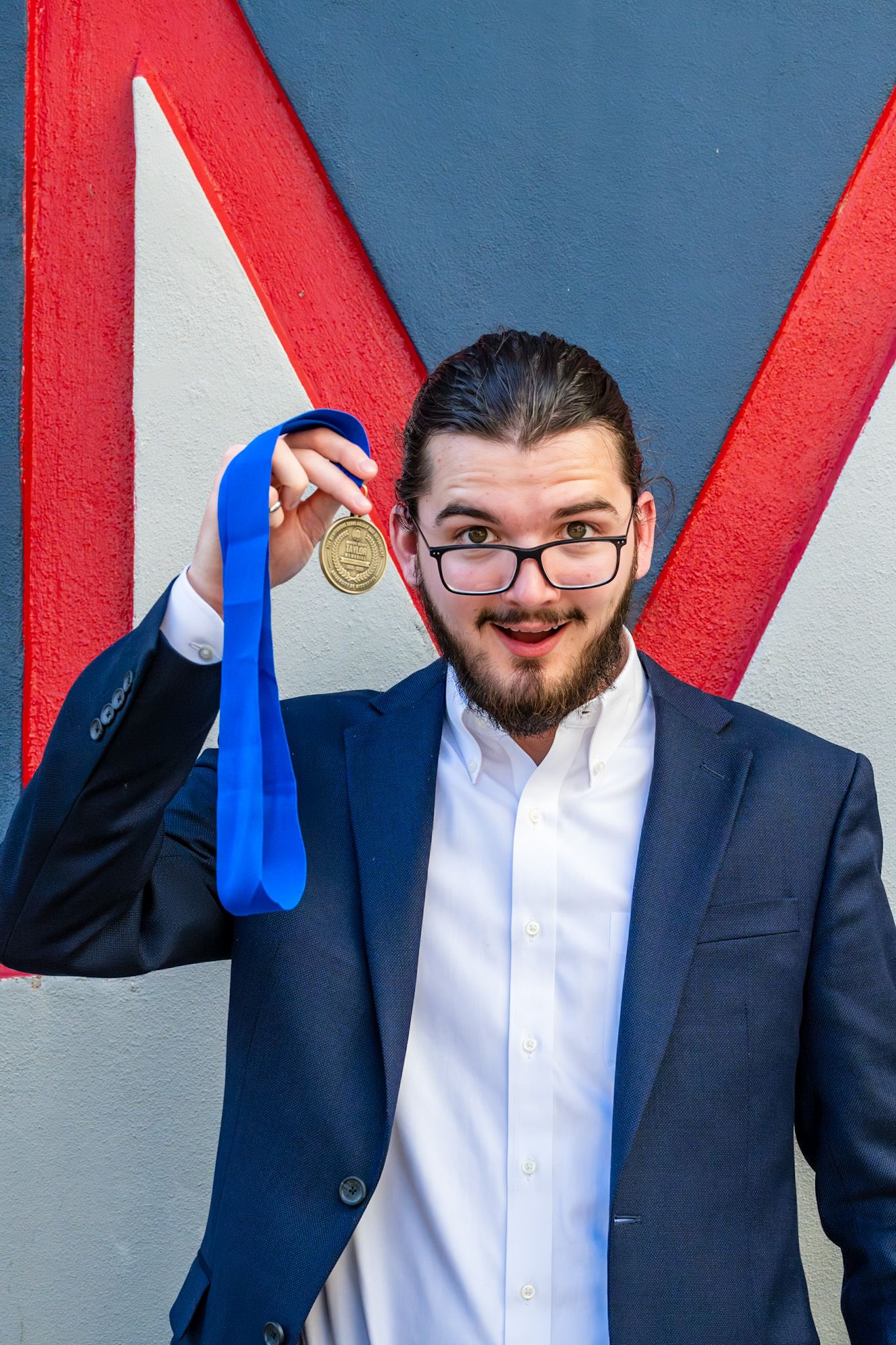 A young bearded man in a navy suit holds up a gold medal with a blue ribbon, smiling proudly against a colorful wall.