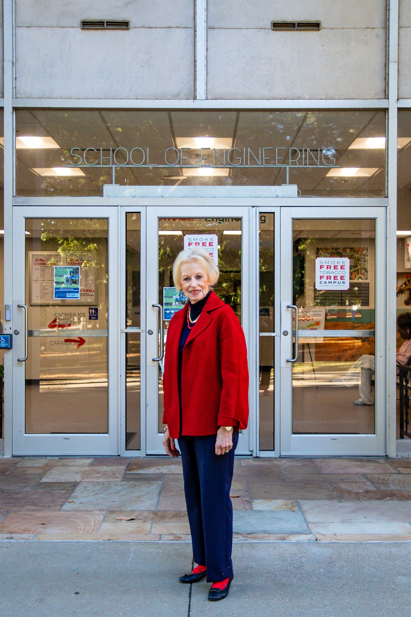 Someone stands at the entrance of a School of Engineering building wearing a bright red blazer and navy slacks.