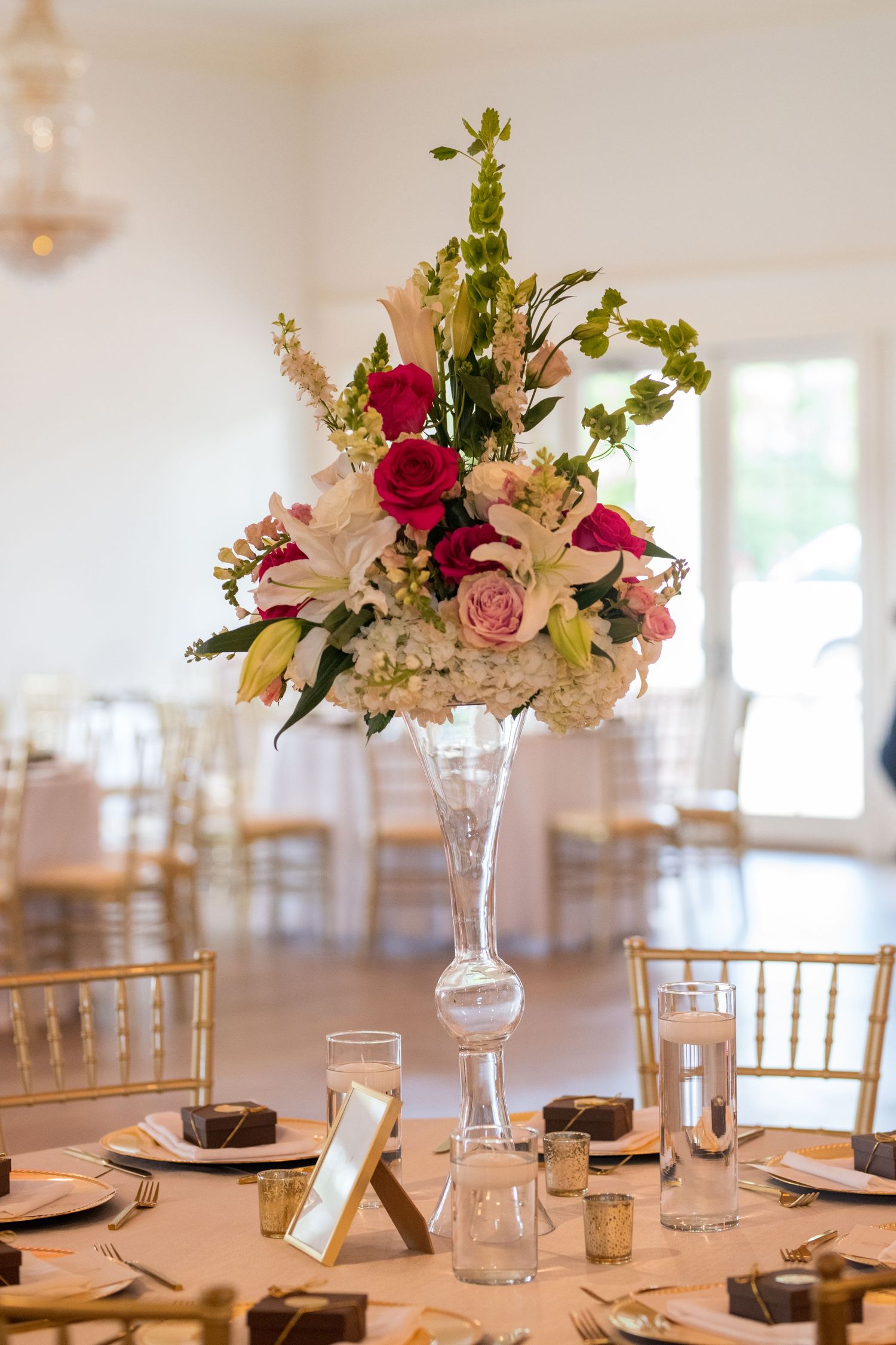 Elegant floral centerpieces with pink and red roses adorn round tables at a formal wedding reception venue setup.