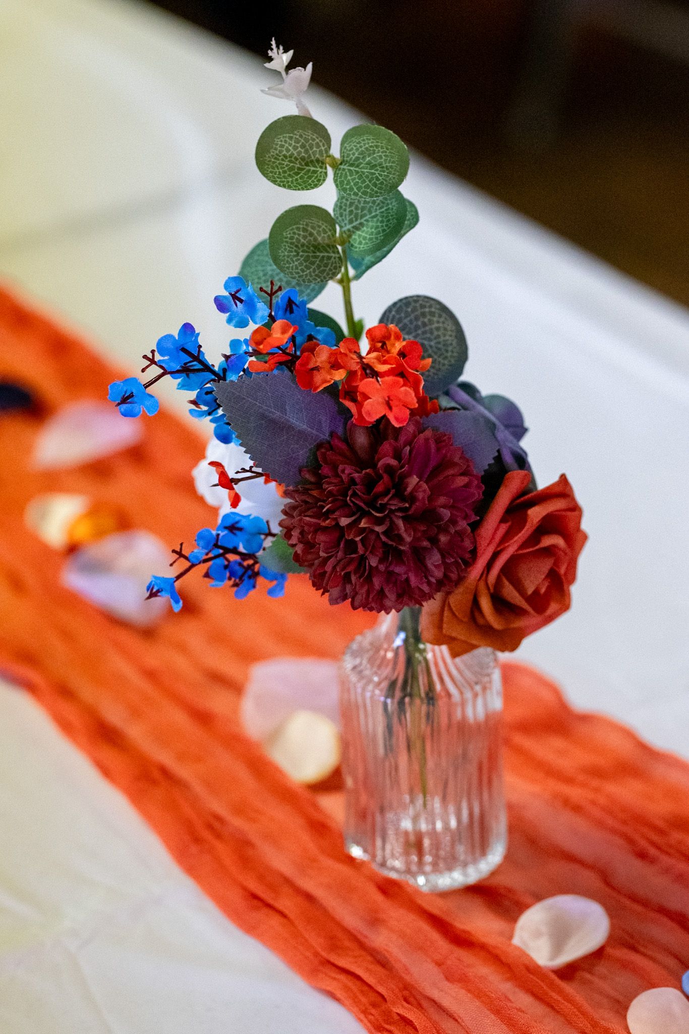 Small floral arrangement with burgundy dahlia, red rose, blue delphinium and eucalyptus in a clear glass vase on orange runner.