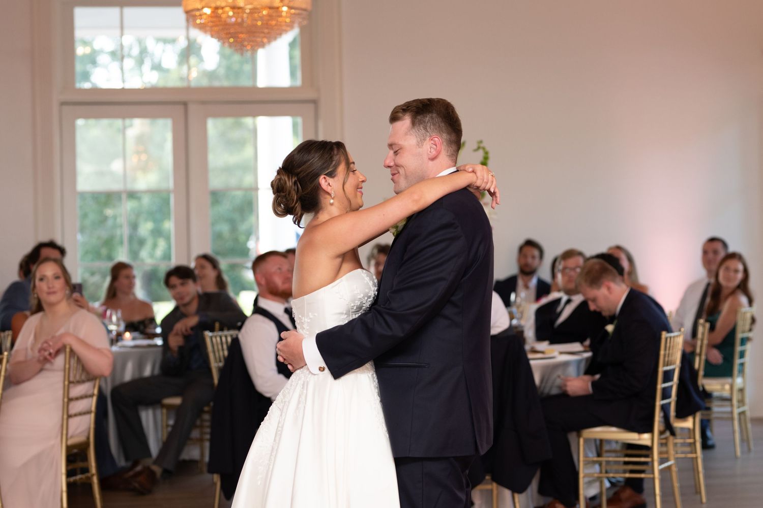 Two people share their first dance as newlyweds at an elegant wedding reception with a crystal chandelier overhead.