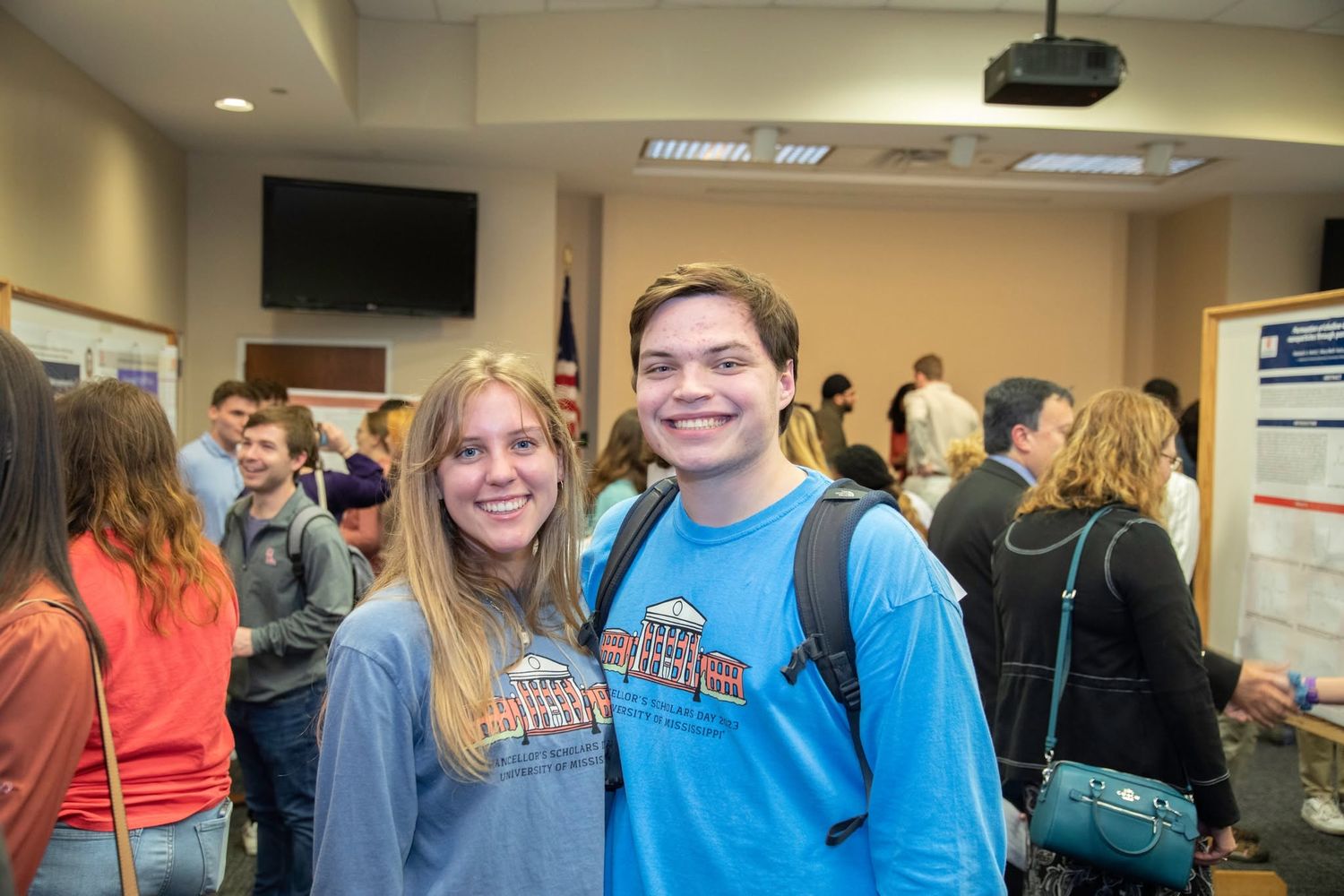 Two smiling college students pose together at an indoor academic event with peers and presentation boards in the background.