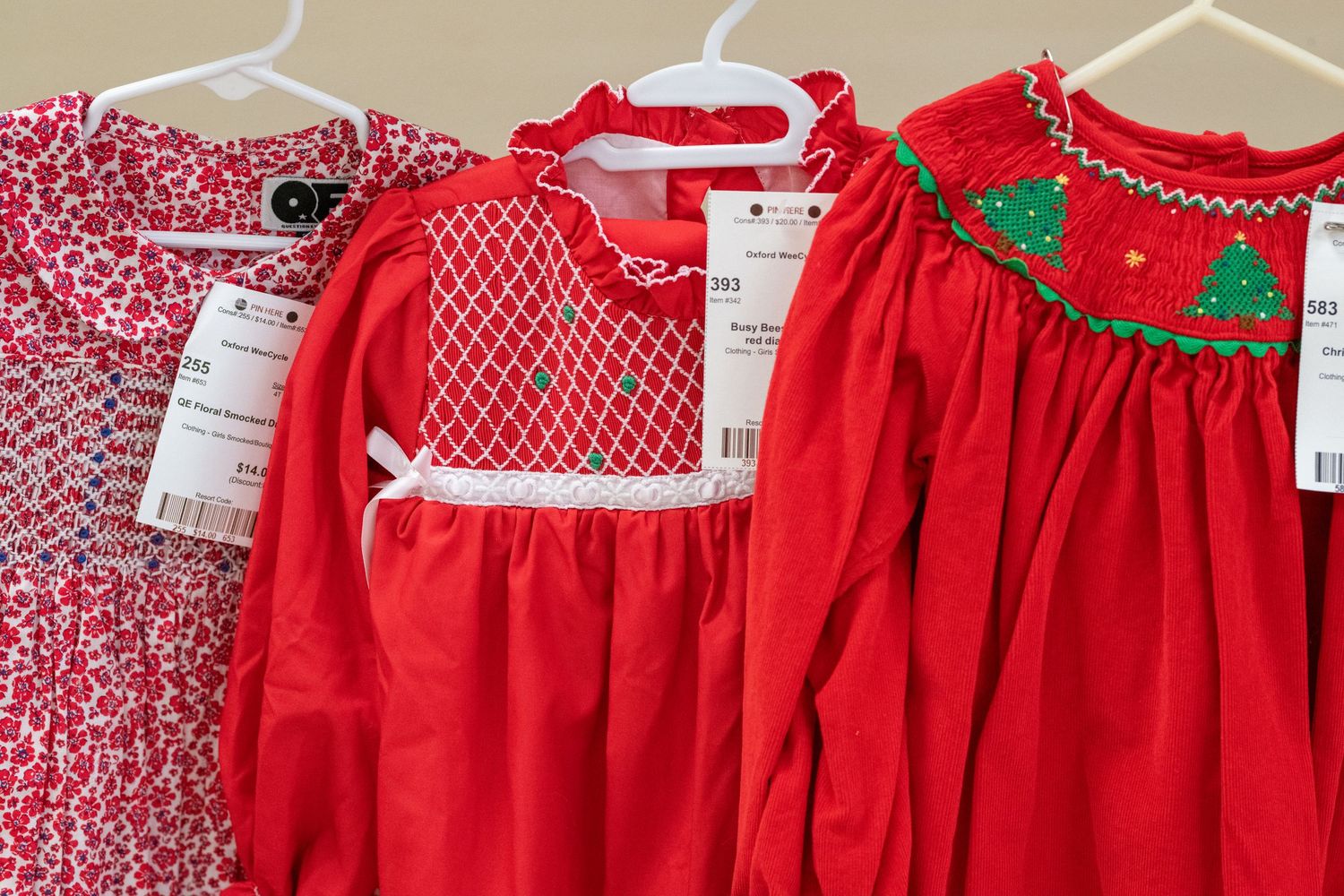 Three red Christmas girls dresses hanging on hangers, including floral, smocked, and Christmas tree embroidered styles.