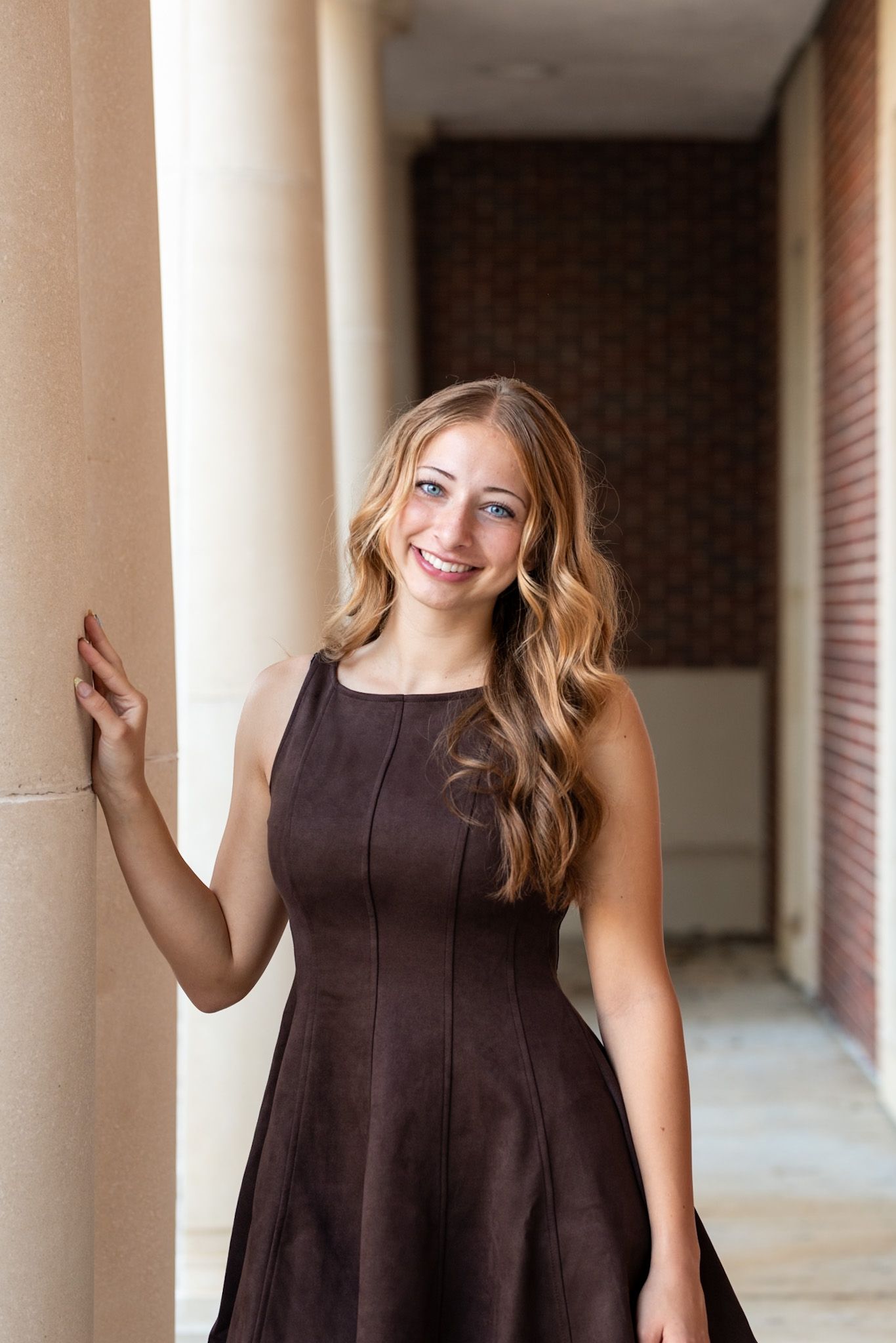 Young blonde woman in brown sleeveless dress smiling while leaning against a column in an elegant hallway.