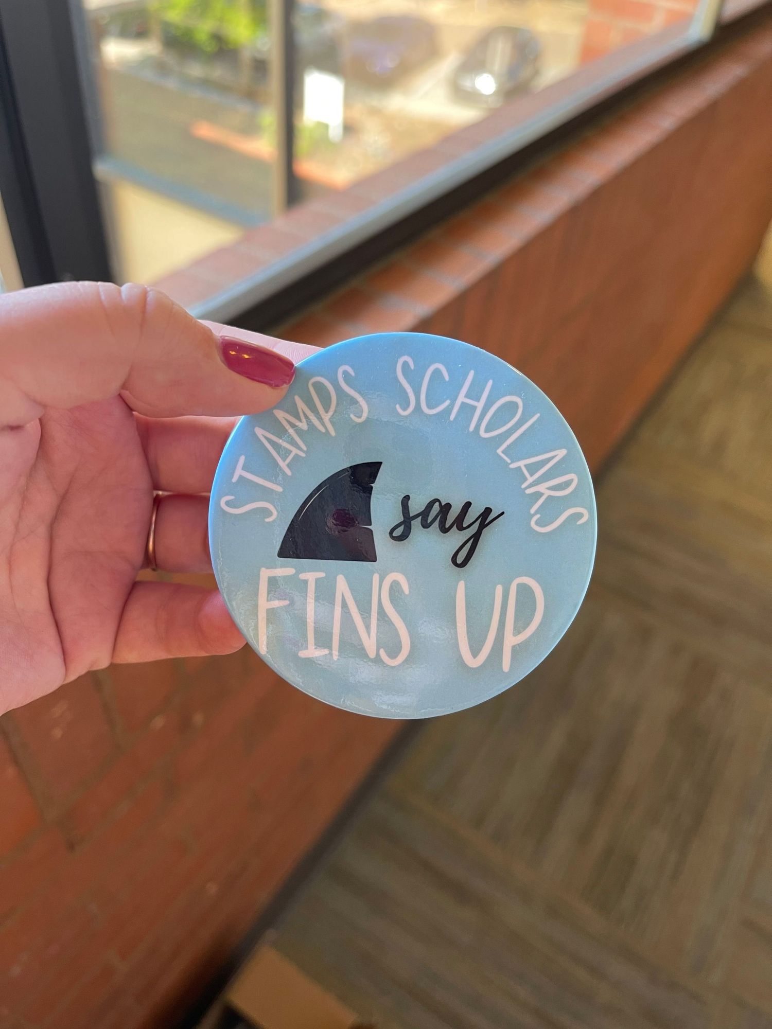 Hand holding a round sticker reading Stamps Scholars Say Fins Up with a shark fin graphic on blue background.