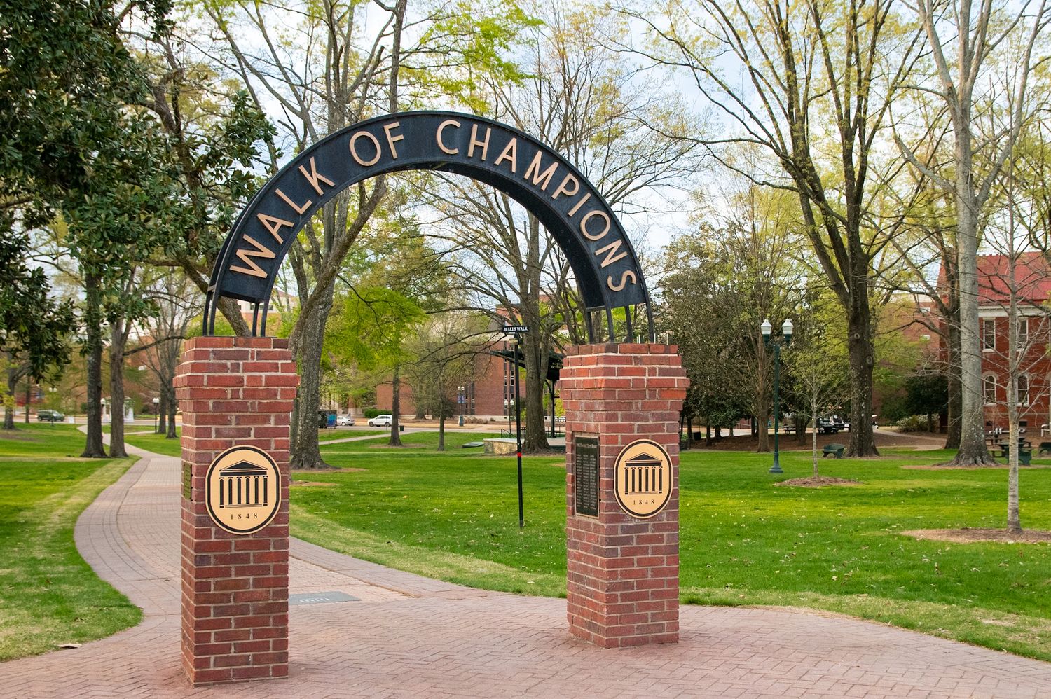 Brick arch gateway reading Walk of Champions on a tree-lined campus path with green lawns.