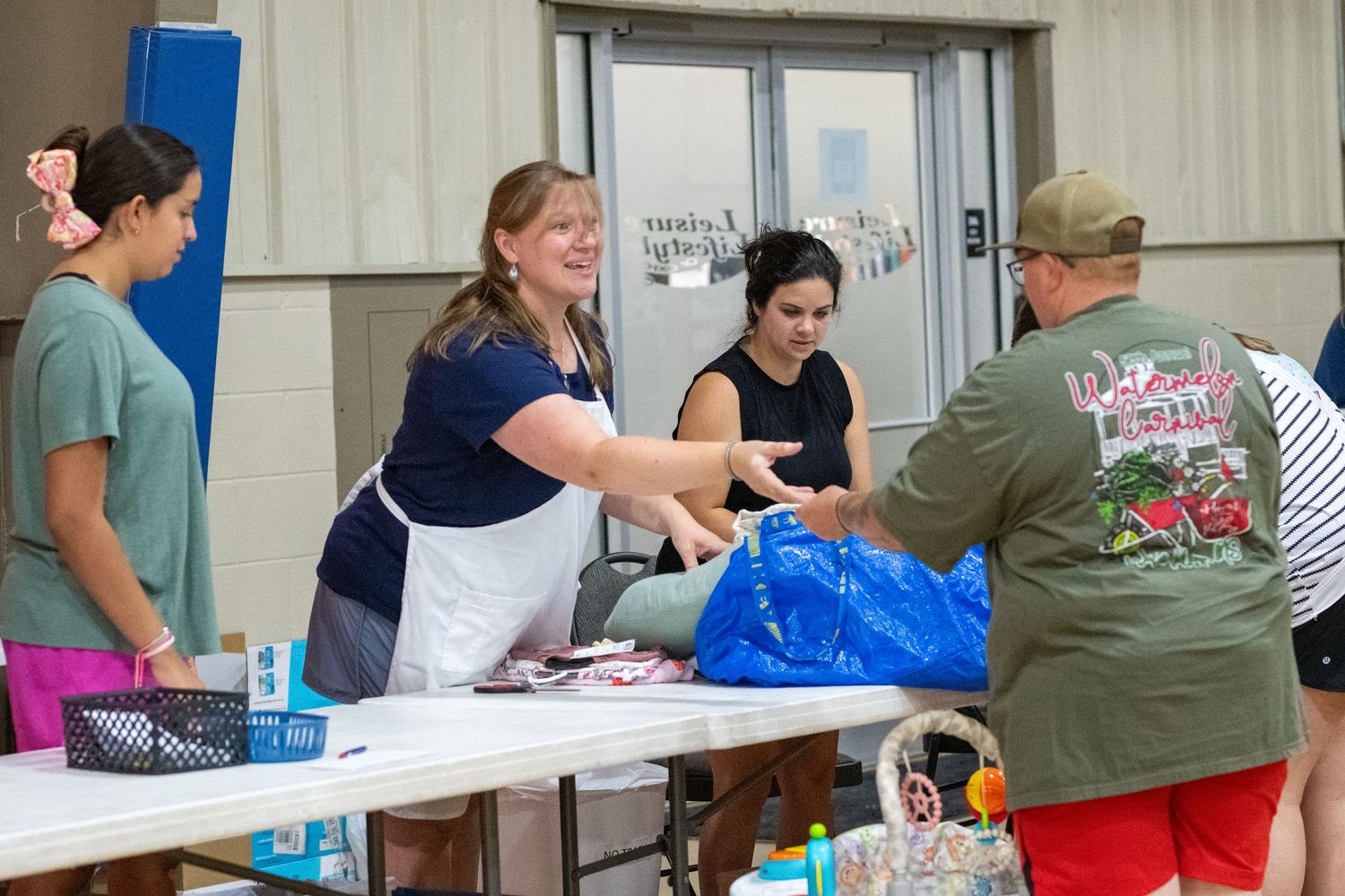 Volunteers distribute supplies at a community aid event, passing bags across a table indoors.