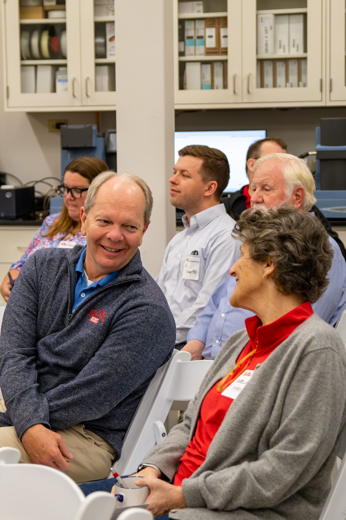 Group of adults seated in a lab setting, smiling and engaged during a professional meeting or seminar.