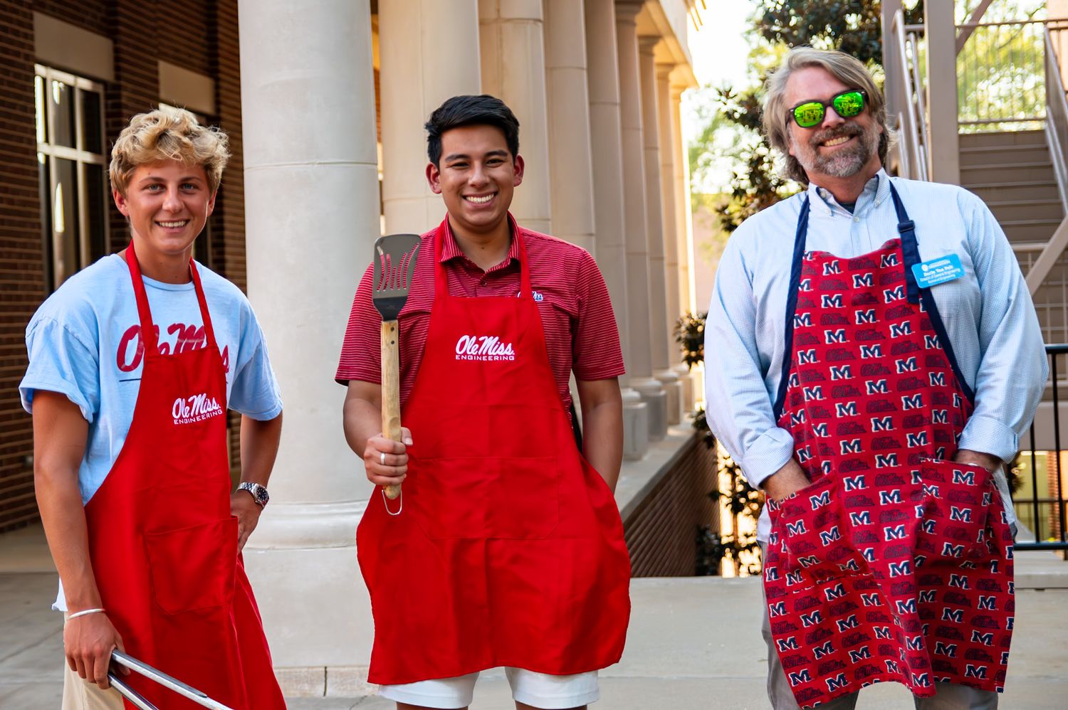 Volunteers in matching red aprons and festive patterned vests stand together in a row at what appears to be a community event.