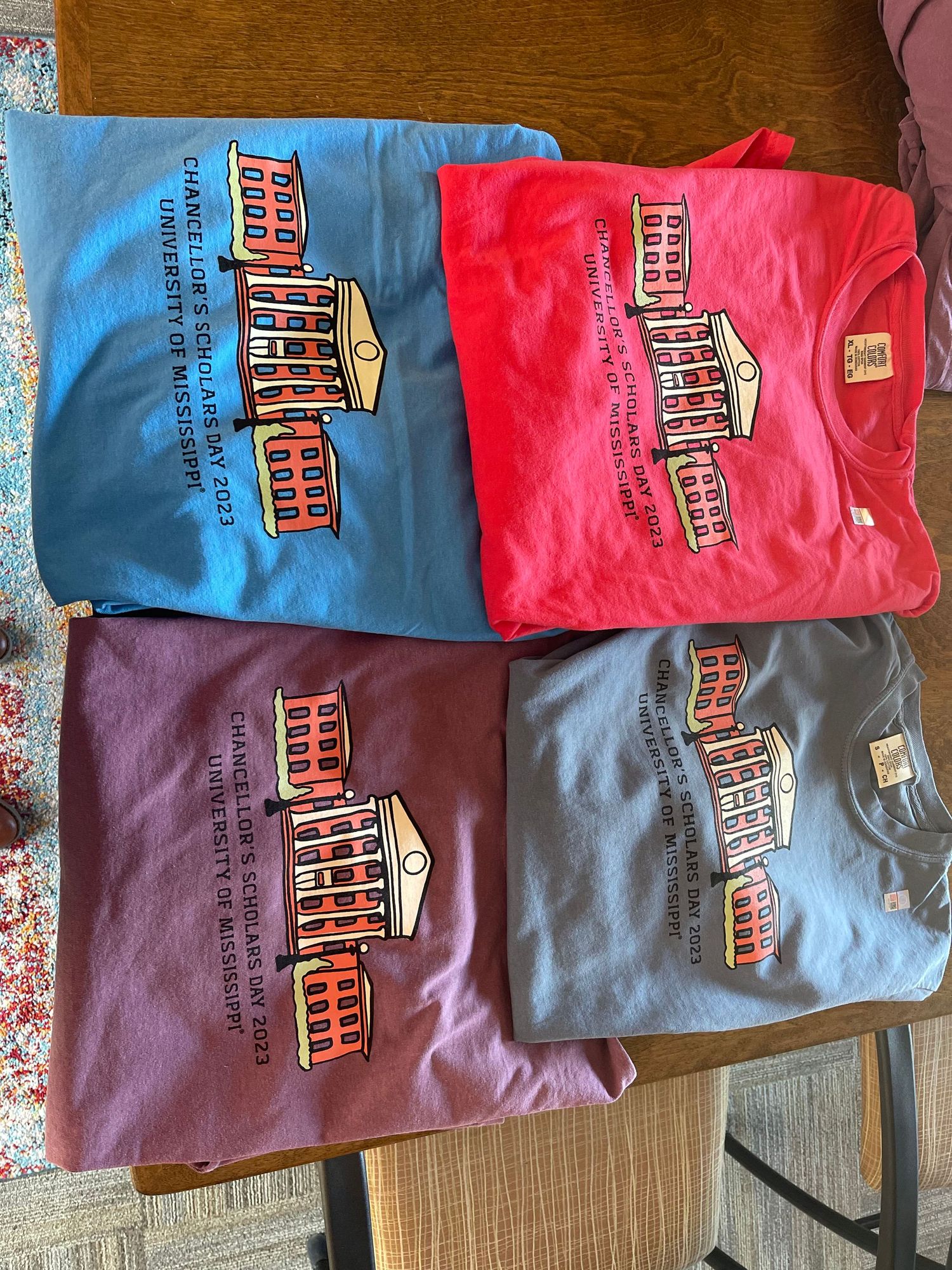 Four colorful University of Mississippi Chancellor's Scholars t-shirts in blue, red, maroon, and gray folded on a table.