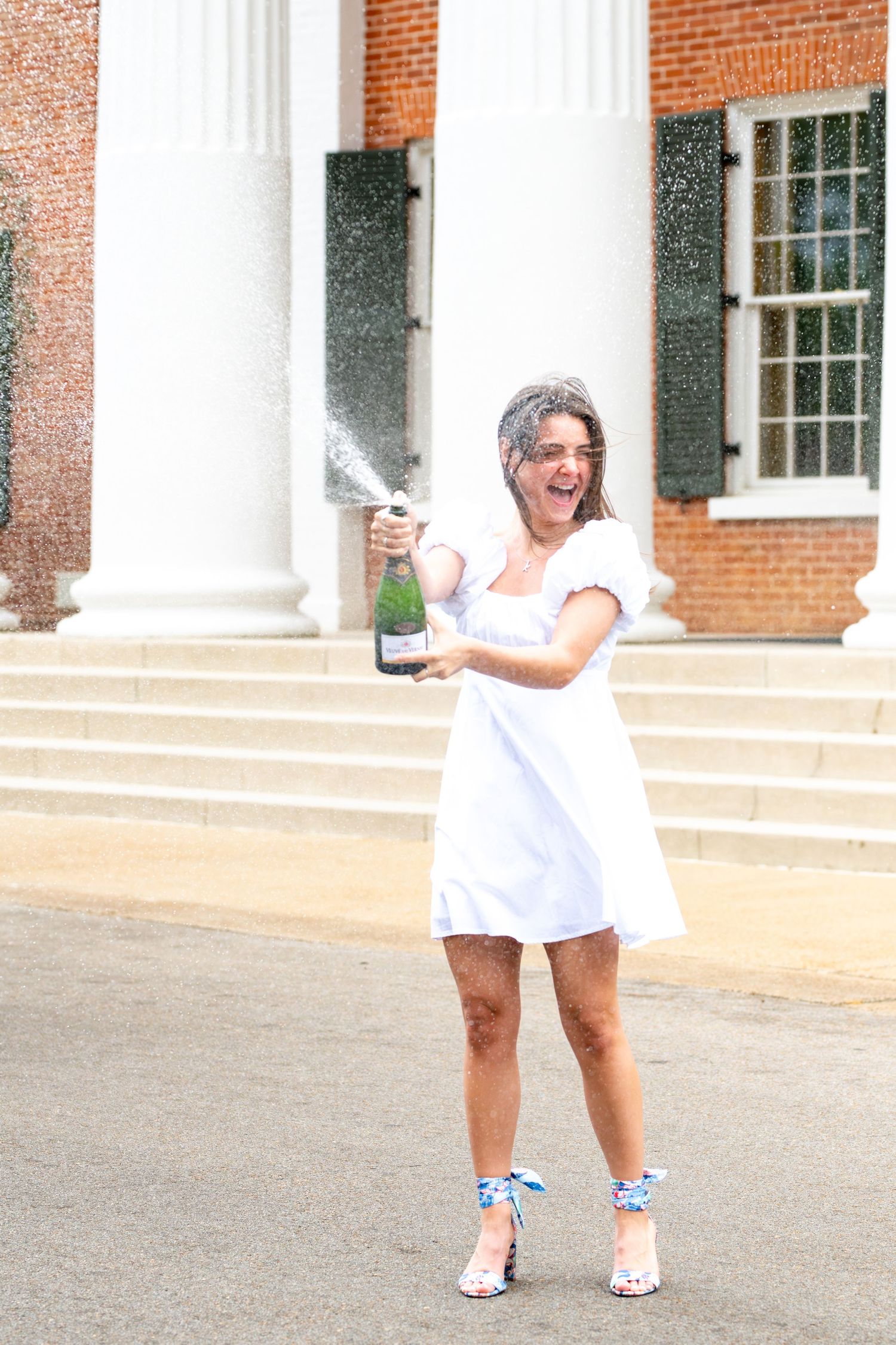 Person in white dress and sparkly sandals celebrates with champagne spray outside brick building with columns.