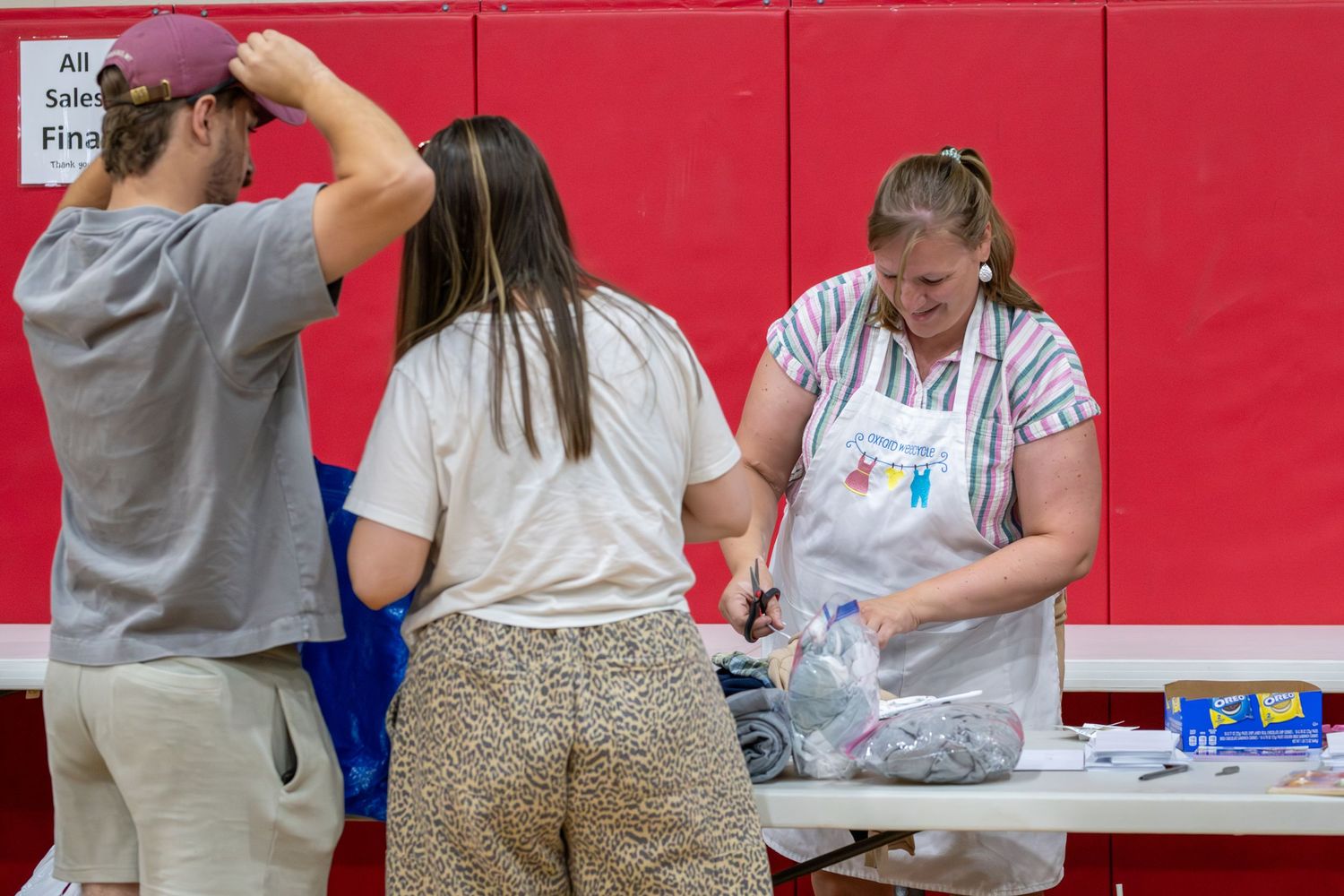 Woman in apron assists customers at a registration table inside a gymnasium with red wall padding.