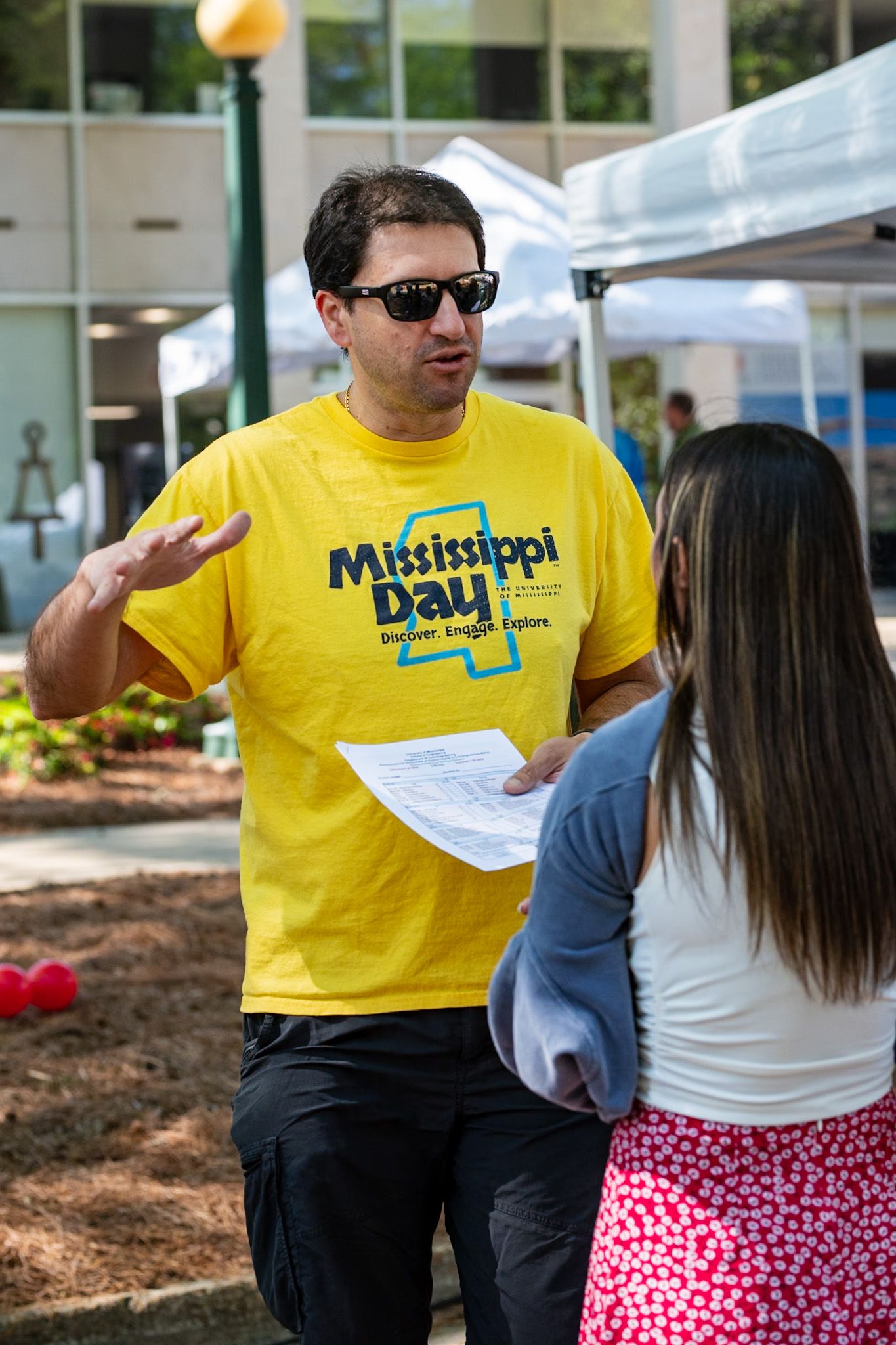 Someone wearing sunglasses and a yellow Mississippi Day t-shirt speaks with another person at an outdoor event.