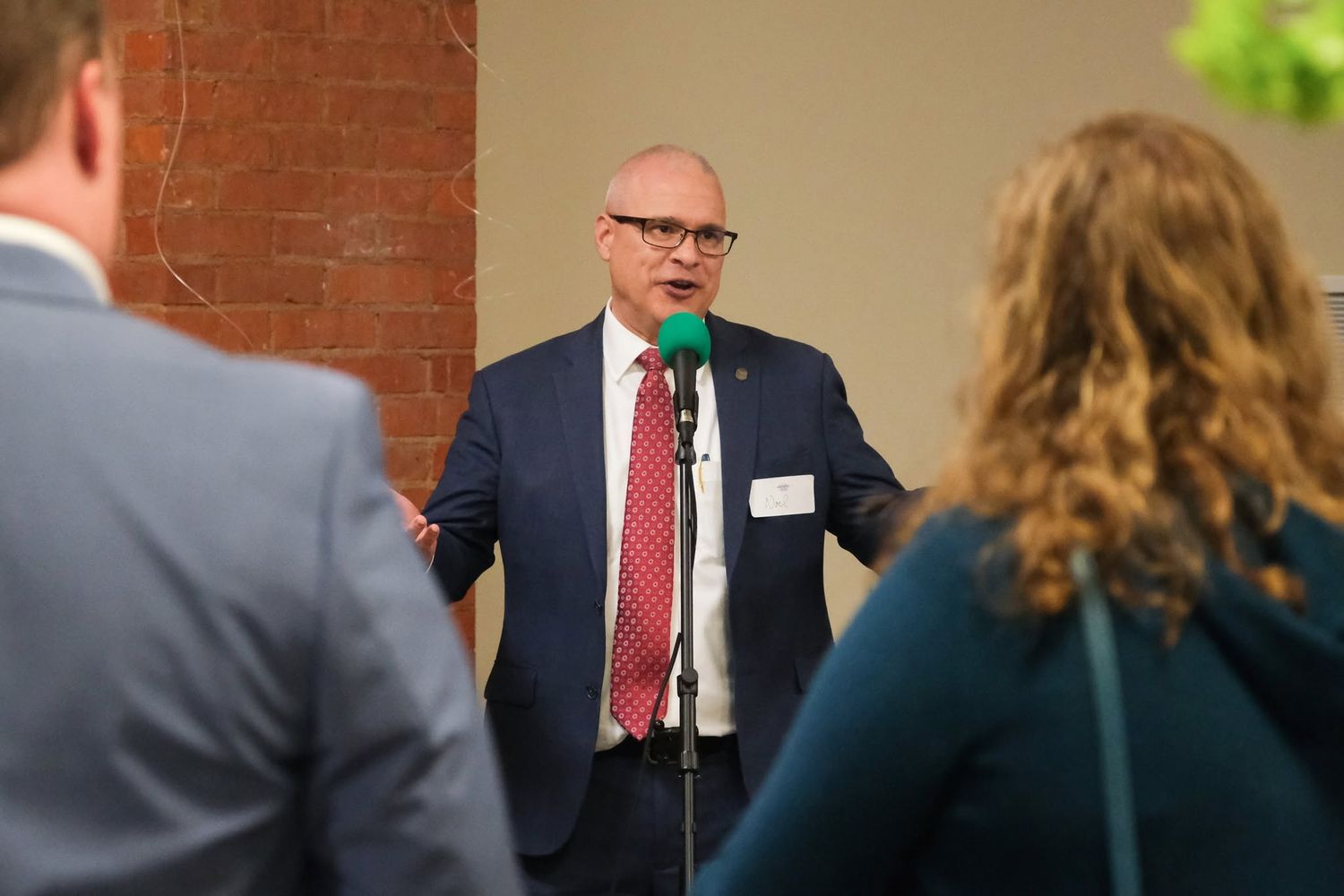 A man in a navy suit and red tie speaks at a microphone during a professional networking event indoors.