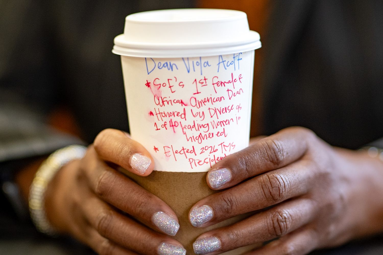 Hands holding a white coffee cup with handwritten notes listing historic achievements in blue and red marker.