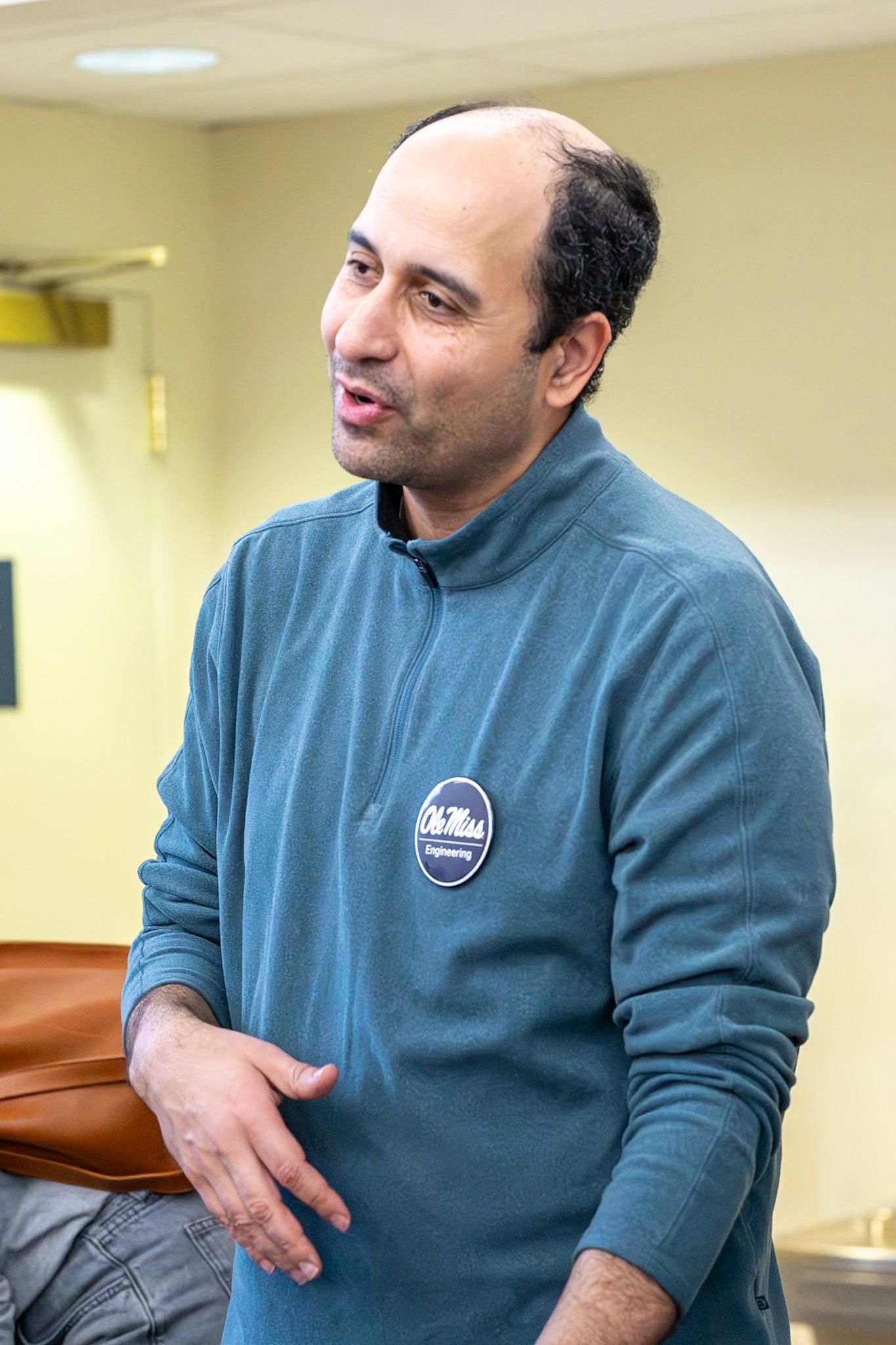 Man in teal Ole Miss Engineering fleece jacket speaking indoors, gesturing with hands in casual setting.