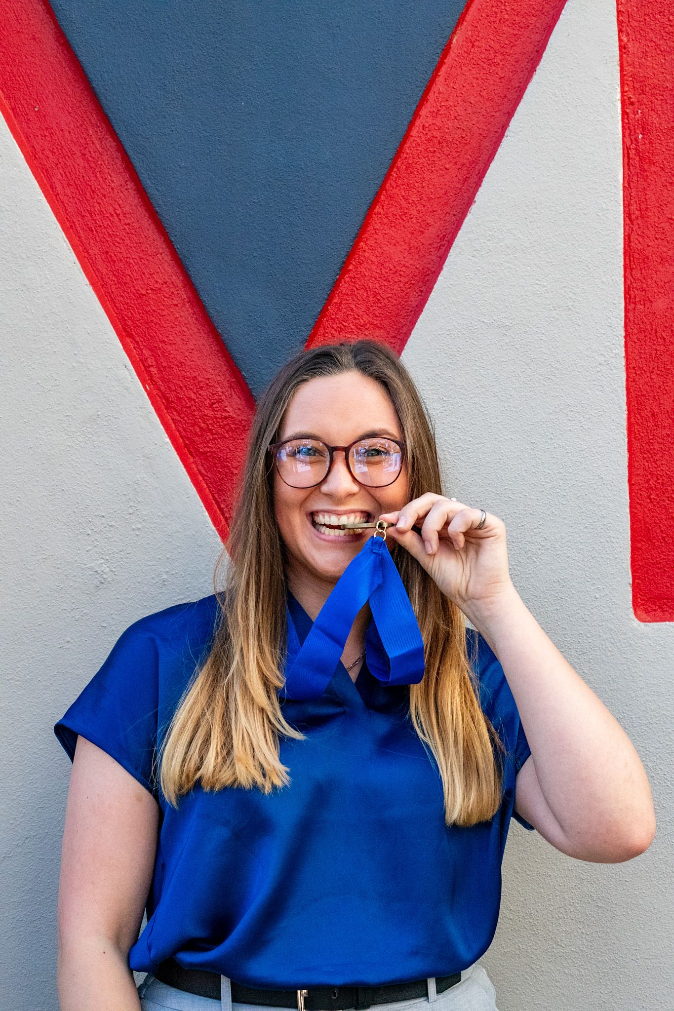 Woman in blue blouse biting a medal with blue ribbon, smiling in front of colorful mural.