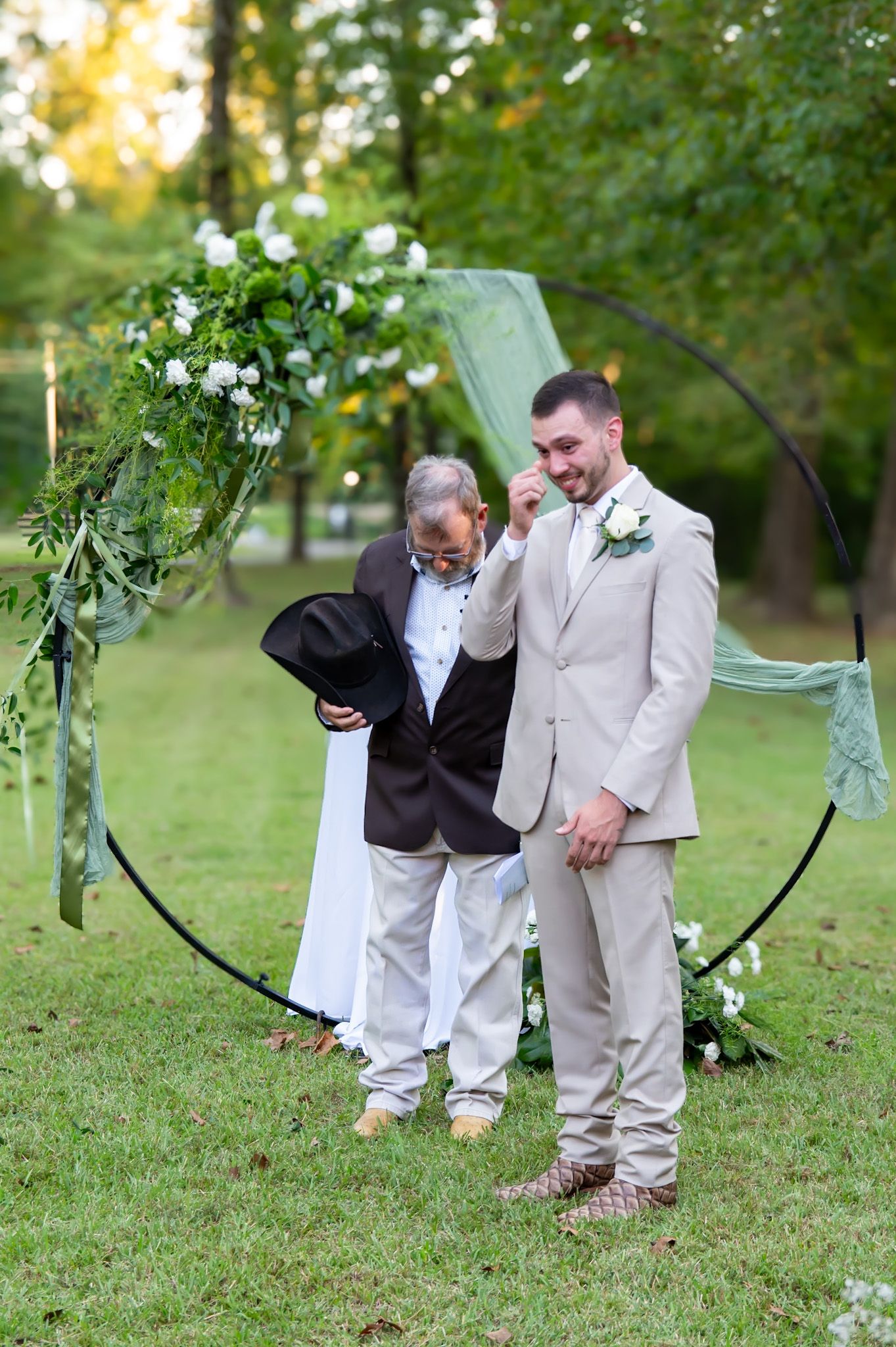 Two people in formal attire laugh and share a candid moment while crouching down in a lush green outdoor setting.