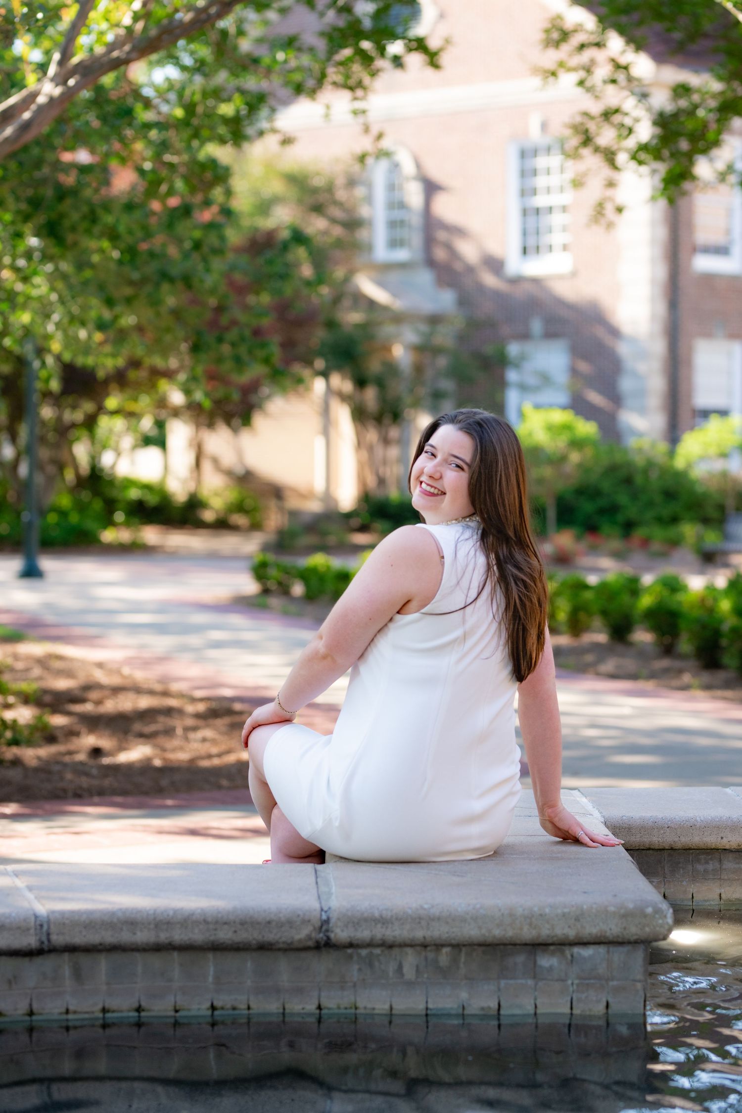 Young woman in white dress sitting by fountain in sunlit garden, smiling over shoulder.