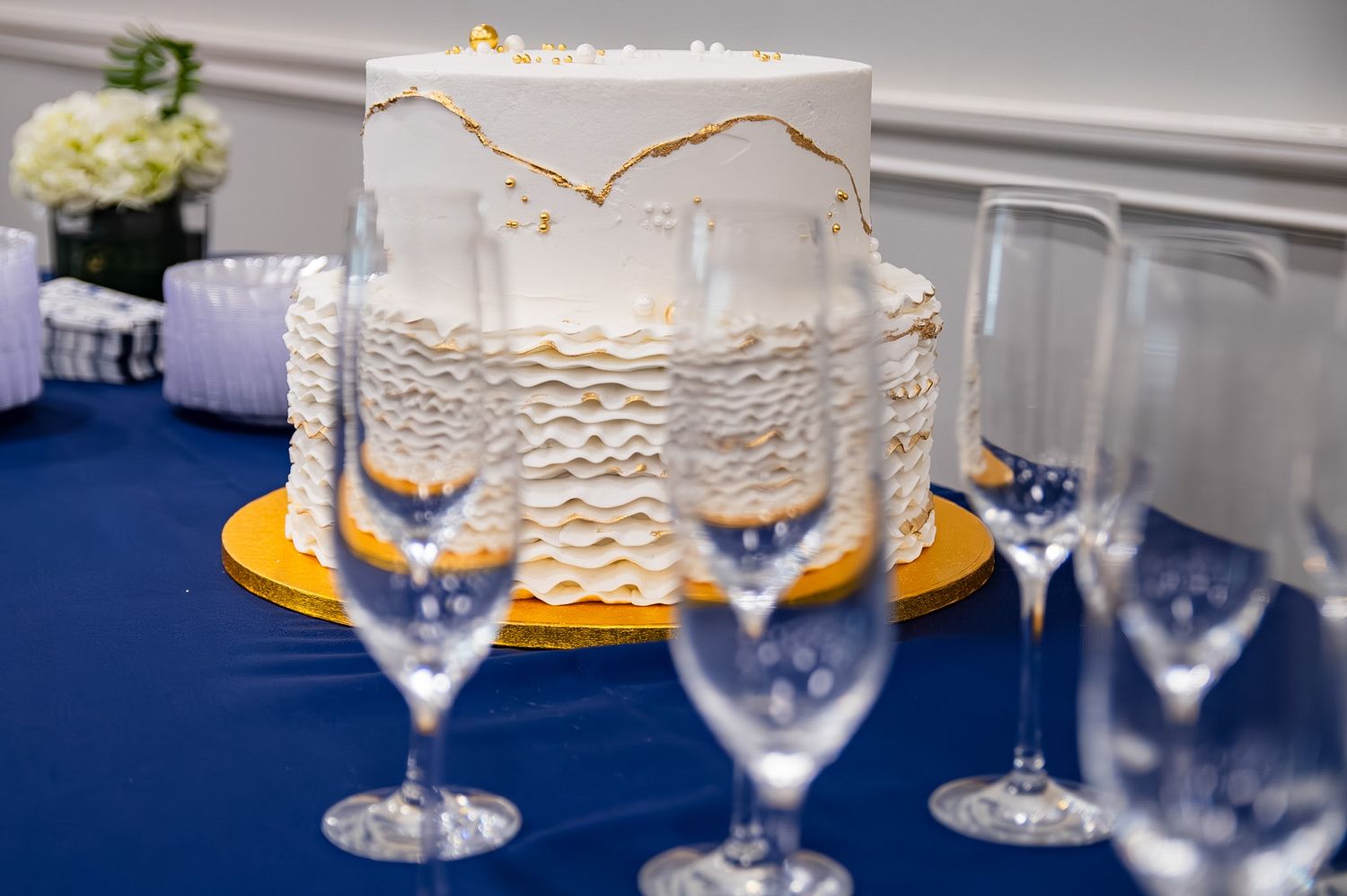 Elegant white wedding cake with gold accents on a blue tablecloth, surrounded by crystal champagne flutes.