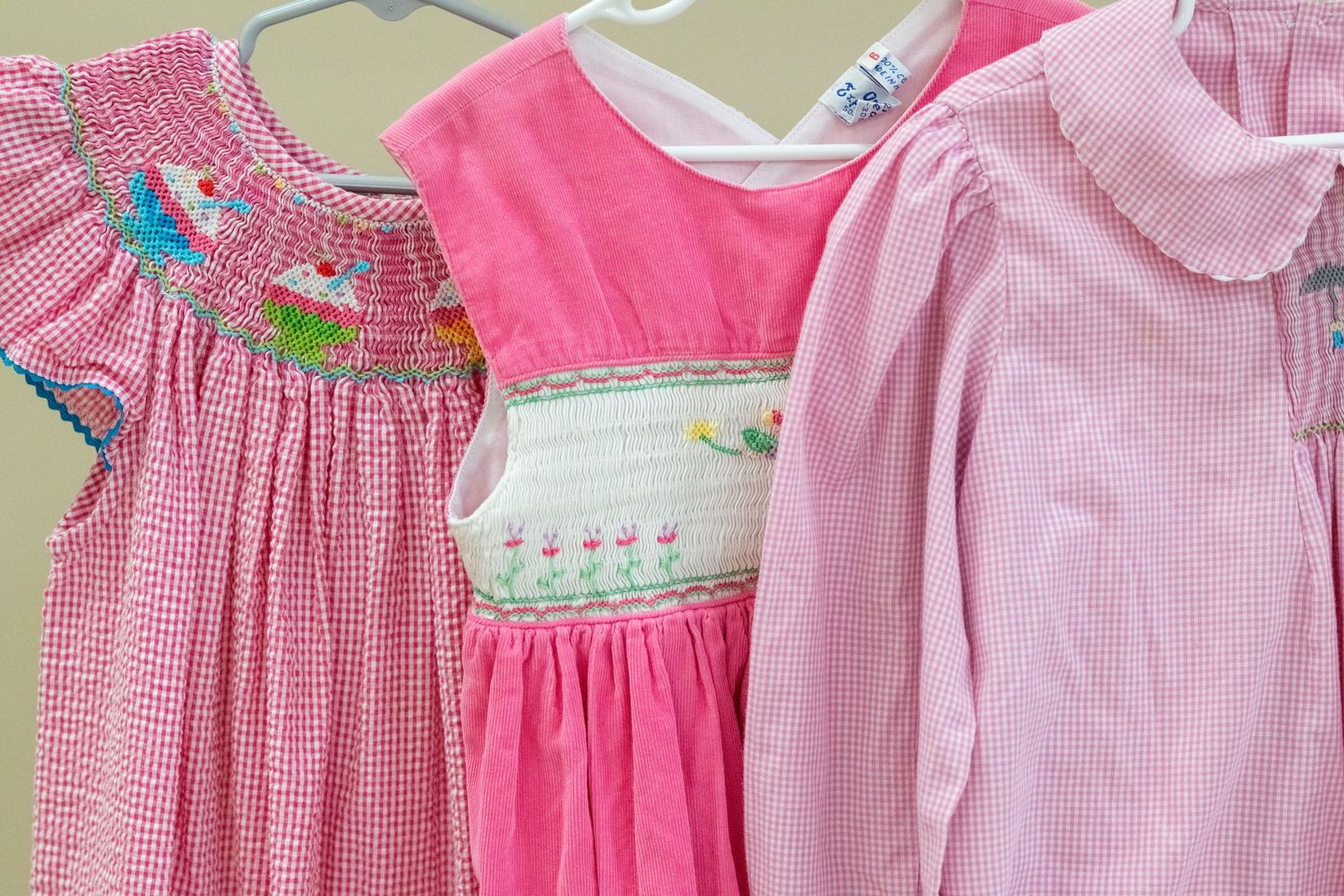 Three pink girls smocked dresses hanging on hangers, featuring ruffles, gingham patterns, and colorful embroidery details.